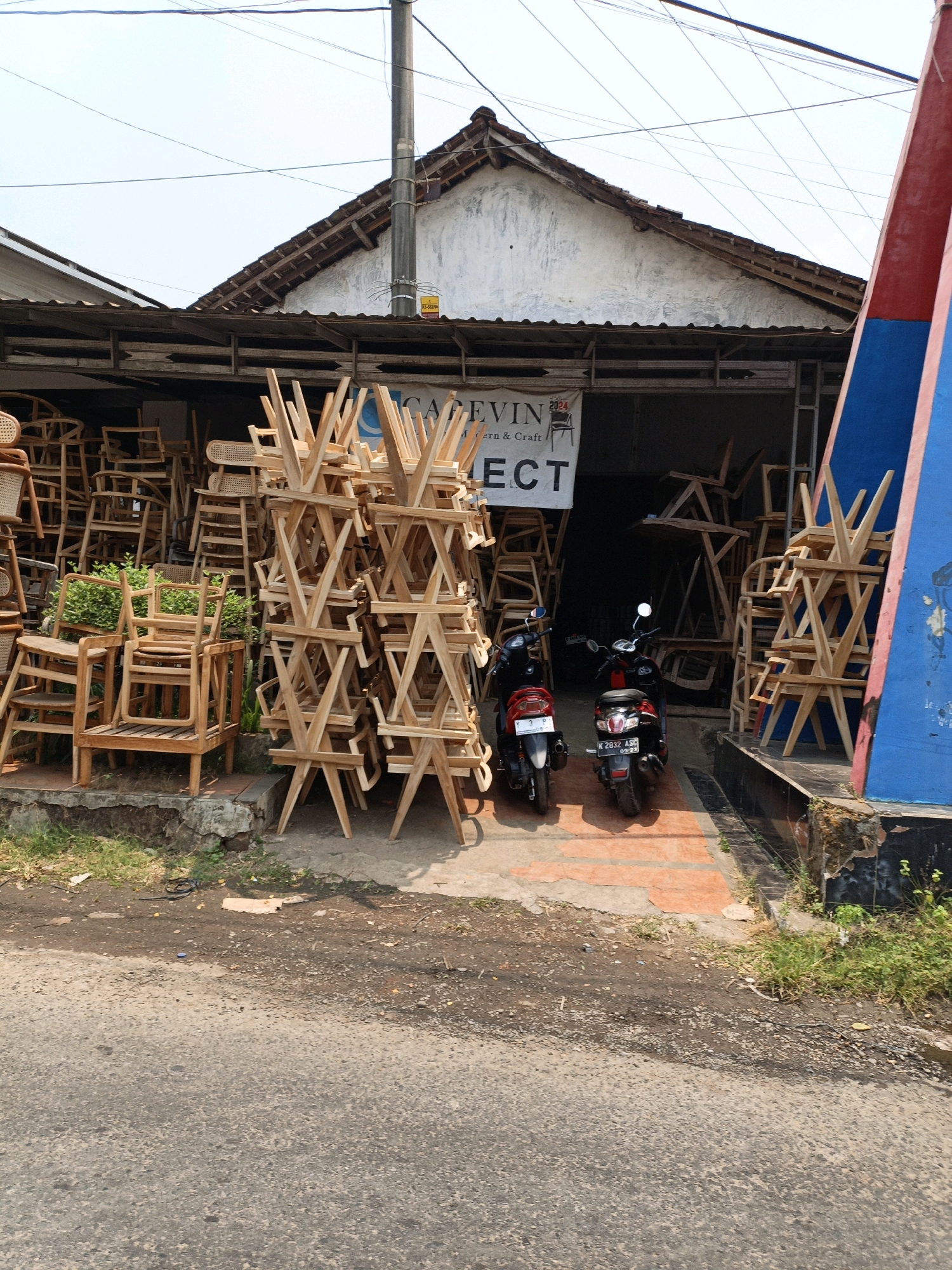 The image captures a storefront in Cikal, Indonesia, specializing in wooden furniture, primarily chairs. The facade is open, revealing stacks of chairs piled both inside and outside the shop. Many chairs are made of wood with woven cane seats and backs, while others are solid wood with unique geometric designs. Two motorcycles, a red and black one and a black one, are parked in front of the entrance. A sign above the entrance reads "KAEVIN" followed by "Modern & Craft" and "LECT". The area is paved in front of the shop, with a dirt and gravel area bordering a grassy patch to the left and right of the entrance. The overall impression is a street-level view of a furniture business in a tropical or sub-tropical environment, given the bright sunlight and the type of vegetation visible.