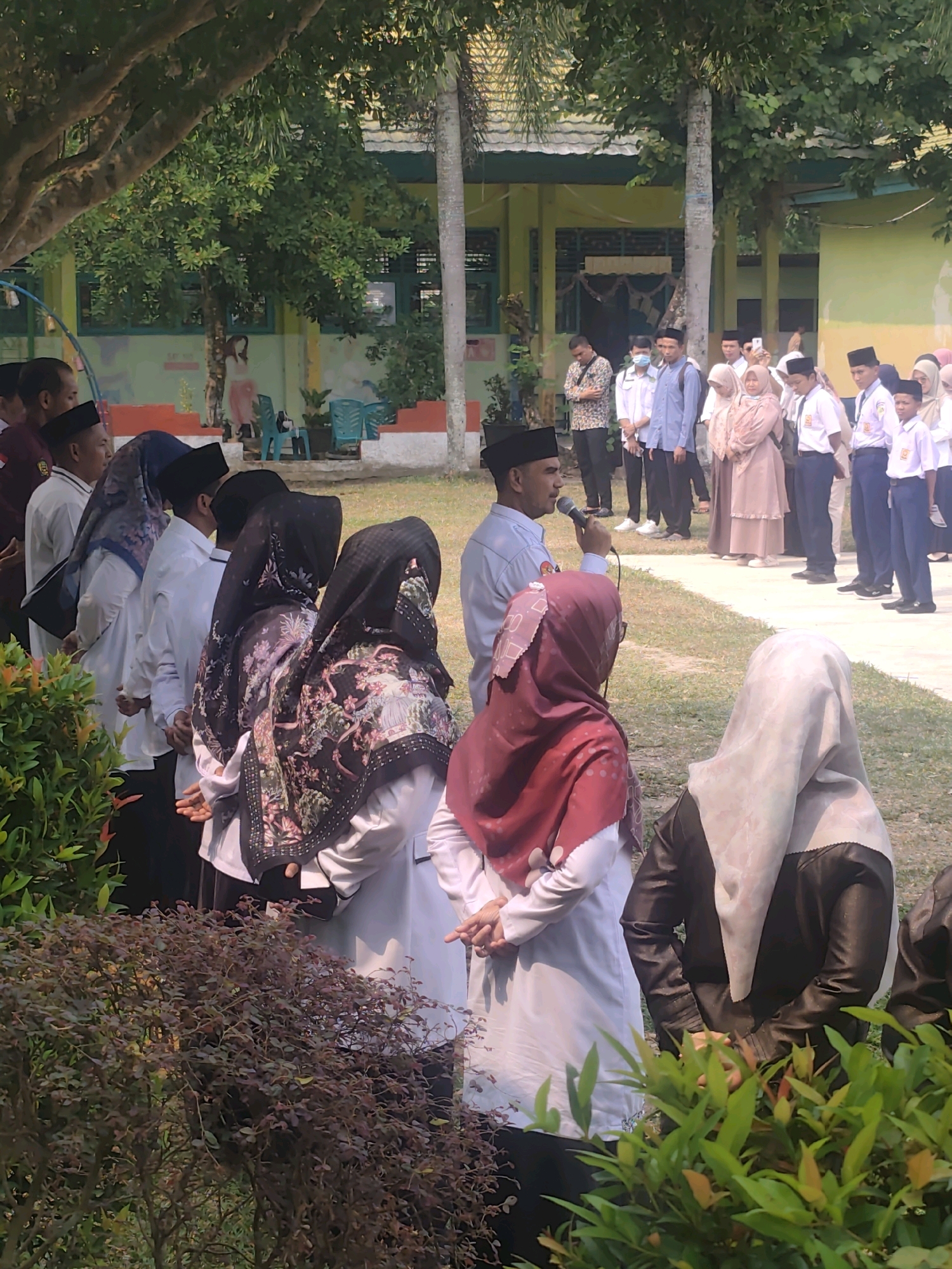 The image depicts an outdoor gathering, likely at a school, in Pematang Reba, Indonesia. In the foreground, several individuals are seen from behind, some wearing hijabs and formal attire. A man in a light blue shirt and black peci, holding a microphone, stands in the middle ground, addressing a group of students. The students, a mix of boys and girls, are dressed in uniforms, including white shirts and blue pants for the boys and various styles of modest clothing for the girls. In the background, a building with a yellow facade and a tiled roof suggests a school environment. Lush green trees and foliage frame the scene, indicating it is daytime.