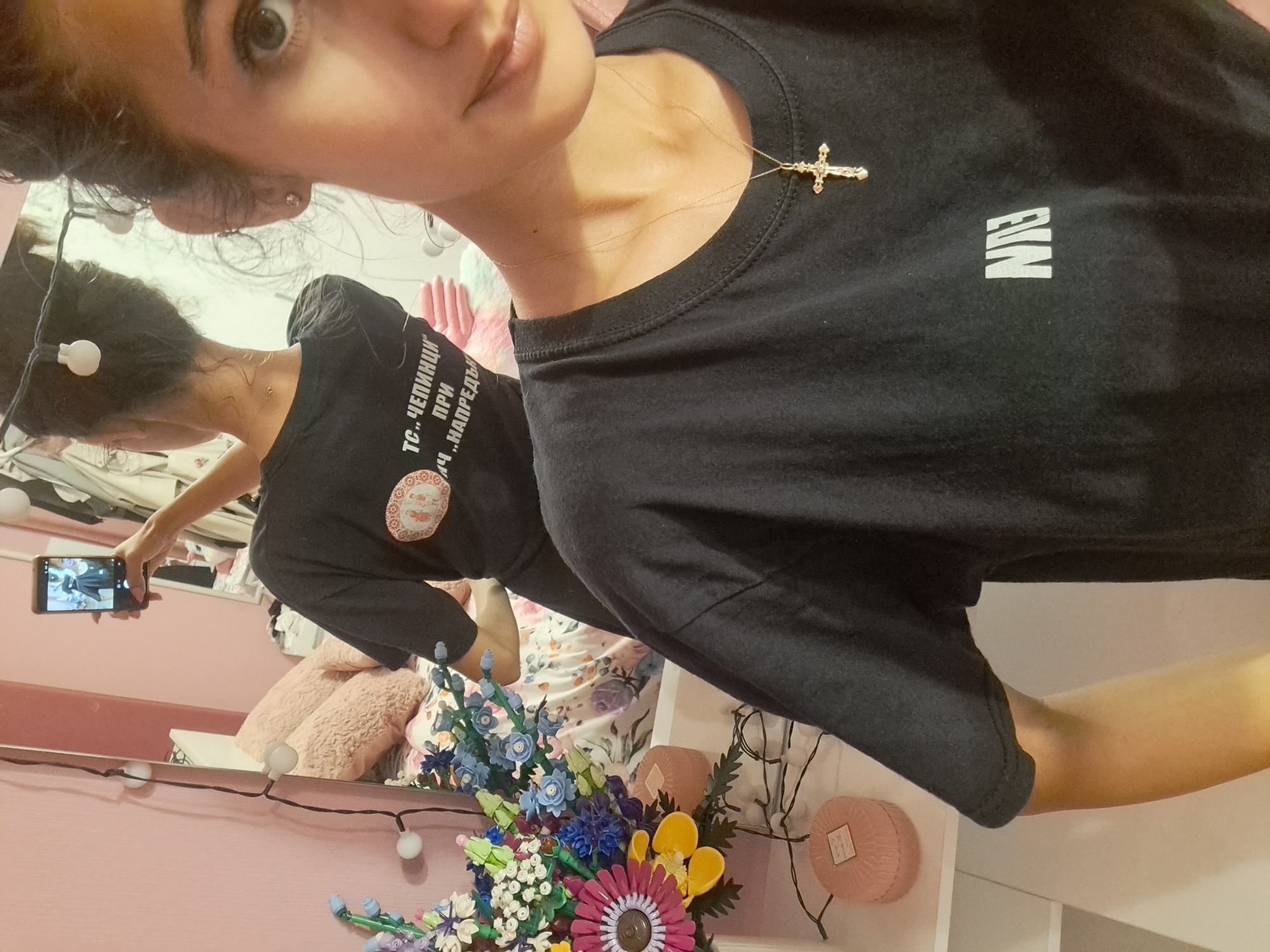 A young woman, viewed from the chest up and to the right, is taking a selfie in a mirror. She is wearing a black t-shirt with a gold cross necklace. The t-shirt has white lettering on the left side and a circular design on the right side, which appears to be two figures within a decorative border. The woman has dark hair pulled back into a bun and is wearing small stud earrings. Her left arm is visible, extended to hold the phone she is using to take the picture. The background shows a bedroom with a pink wall, a mirror, a bed with pink and floral bedding, and a shelf with decorative items including a bouquet of colorful artificial flowers and two round, pink objects. String lights with white bulbs are draped across the mirror and wall.