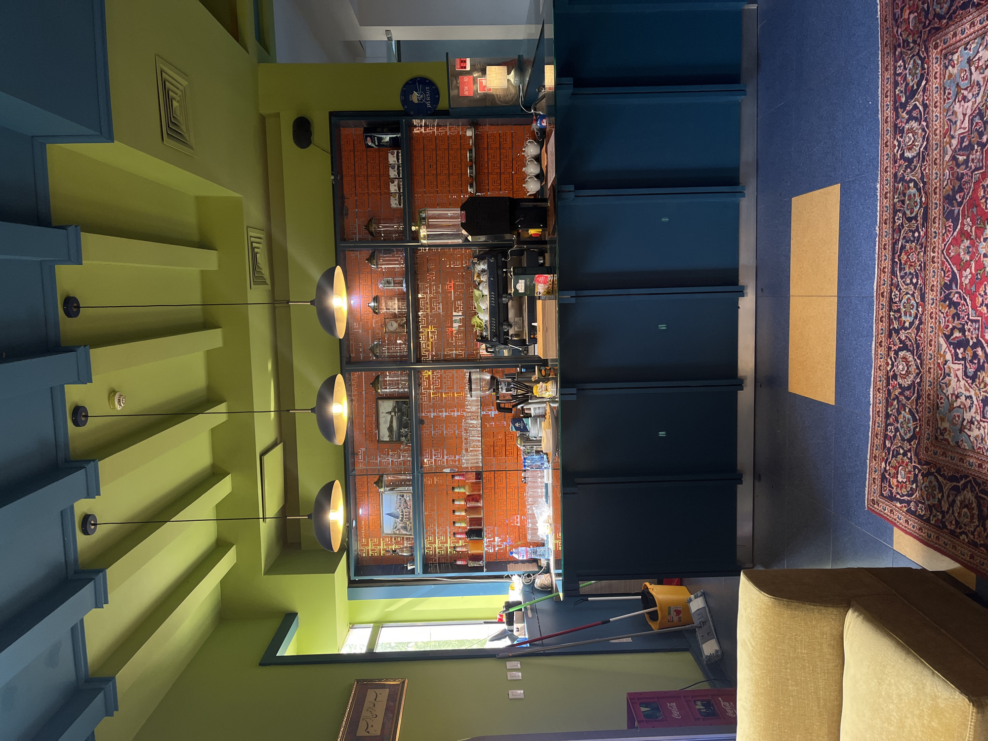 This media file captures an interior shot of an eclectic cafe, bar, or lounge area, likely located in Plovdiv, Bulgaria. No people are visible, suggesting the space is empty or awaiting customers. The vibrant decor features lime green walls and ceiling, accented by dark teal architectural beams and structures.
The central focus is a well-stocked bar counter area. Behind a dark teal counter, a patterned orange and red back wall, possibly tiled or wallpapered, serves as a backdrop for shelves filled with various glasses, bottles, and jars. Bar equipment includes a coffee machine, large clear dispensers, and small white teapots neatly arranged on the counter. A blue circular clock with "PERSUIT" written on its face hangs above, and a red digital display shows "8 / PAC-10".
The floor features dark blue tiling or carpeting, partially covered by a rectangular light brown mat and a large, ornate red and blue patterned rug. In the bottom right corner, a section of a light brown upholstered armchair or sofa is visible. Cleaning supplies, including a yellow bucket and a mop, are seen near a window in the lower left, through which some outdoor light and greenery are visible. A red "Coca-Cola" crate sits on the floor nearby. Further along the lower wall, there's a framed picture containing what appears to be Arabic calligraphy. The ceiling is fitted with square air vents and illuminated by multiple modern pendant lights with warm glows. The time of day appears to be daytime, with natural light entering through the window, complemented by interior lighting.