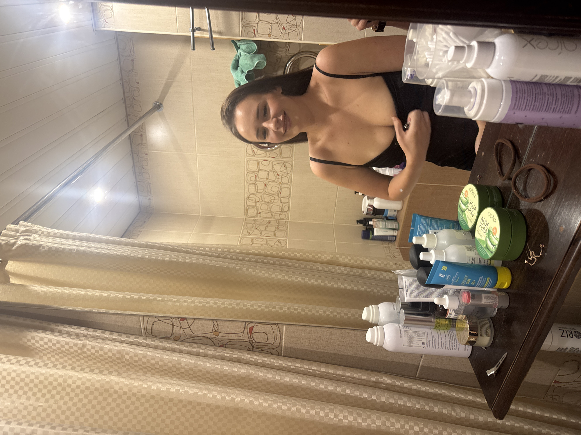 A woman is taking a selfie in a bathroom. She is smiling and looking at the camera, and she is wearing a black top. In the background, there are toiletries on a shelf, including bottles of lotion and shampoo. There is also a towel hanging on the wall. The room appears to be a typical bathroom, with tiled walls and a shower curtain. The lighting is bright, suggesting it is daytime. The overall impression is one of a candid, everyday moment.