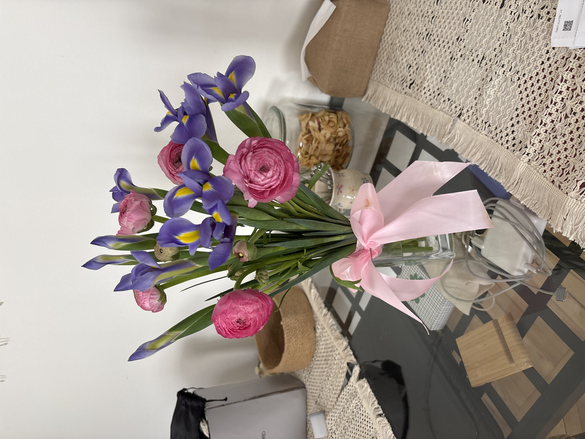 A bouquet of blue and purple irises and pink ranunculus flowers, tied with a light pink ribbon, sits on a glass-topped table with a patterned surface. The table is covered with a light-colored, fringed tablecloth. Next to the bouquet is a jar of what appears to be dried apples. A white wall is partially visible on the left, and various objects, including a shopping bag and electronic cables, are scattered around the table. The location in Moscow, Russia is not determinable from the image.