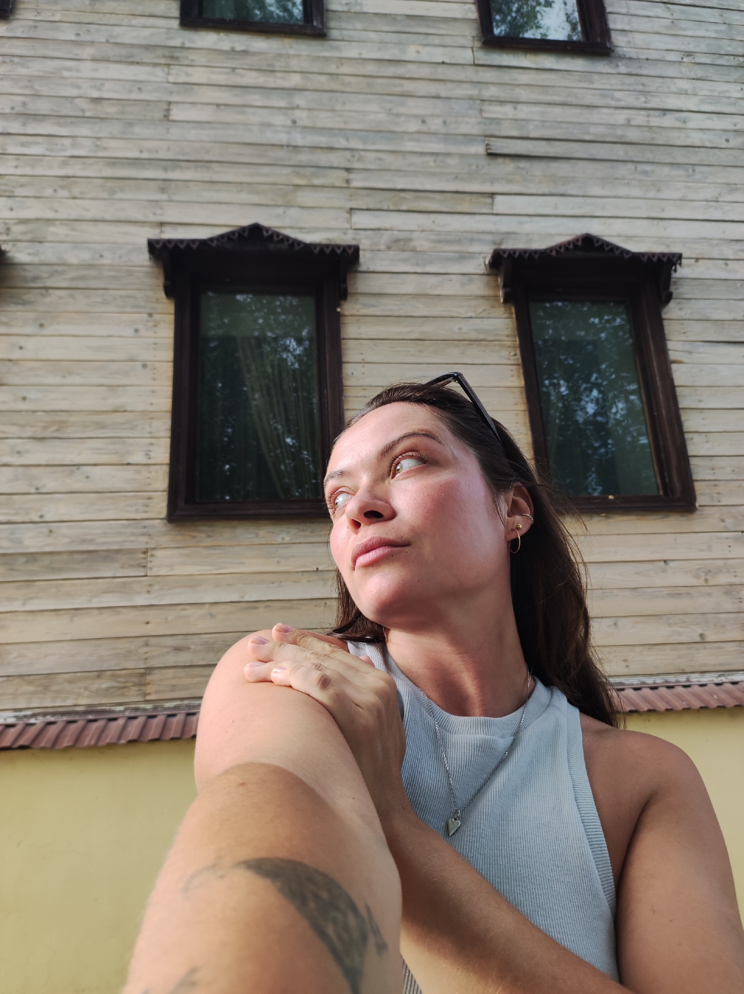 The image shows a woman posing in front of a wooden building with several dark-framed windows. She is looking upwards and to her left, with her right arm extended across her chest and her hand resting on her left shoulder. She is wearing a light blue tank top and a silver pendant necklace. There is a tattoo visible on her left arm. The building's exterior is constructed of horizontal wooden planks, and there appears to be a yellow wall and a dark, tiled roof visible below the wooden structure. The setting is likely outdoors, given the natural lighting and the reflection of trees in the windows. The specific location in Zavod Mikhailovskii, Russia, is not evident from the image itself, but the architectural style of the building, particularly the decorative elements above the windows, might suggest a regional influence.