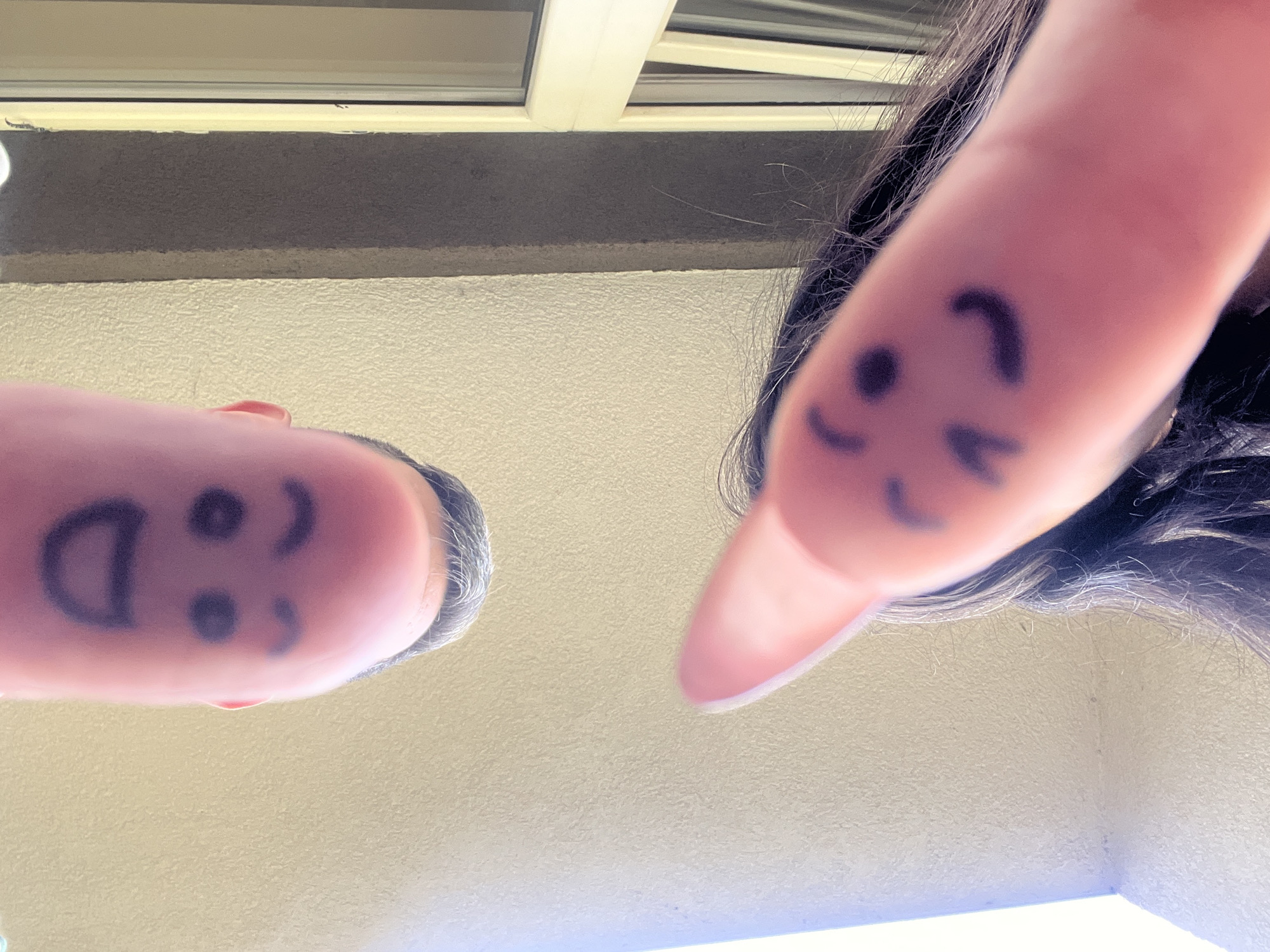 Two fingers are the primary subjects of this close-up, upward-facing shot. On the left finger, a smiley face is drawn with a marker, featuring a "D" for a mouth, two dots for eyes, and a comma for a nose. The finger appears to be attached to a person with short, graying hair, and the back of their head and ear are visible. On the right, another finger is shown with a simple smiley face drawn on it, consisting of two curved lines for eyes and a crescent shape for a mouth. This finger belongs to a person with long, dark hair that is partially obscuring the finger and face.
The scene appears to be outdoors, with a textured wall in the foreground and a white structure with horizontal beams visible in the background. A hint of what might be a window or overhang is present at the top. The lighting suggests it is daytime, possibly late morning or afternoon, as there is bright, diffused light. The overall mood conveyed by the drawn smiley faces is cheerful and playful. No text is visible in the image. The setting appears to be a residential or public area with architectural elements.