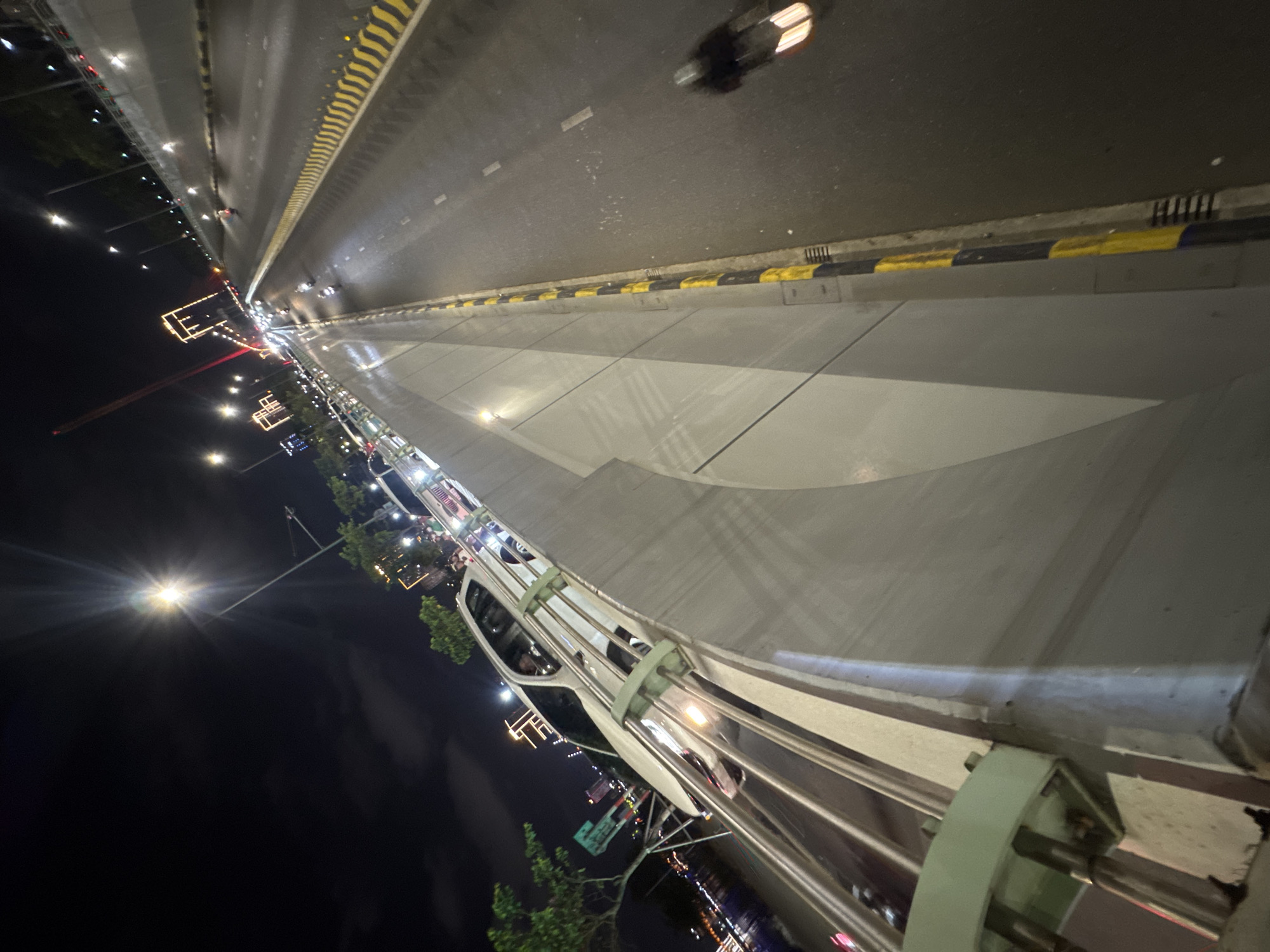 This image depicts a night scene from a bridge in Đà Nẵng, Vietnam, angled in a perspective that skews the horizon. The top of the frame shows a multi-lane roadway with yellow and black striped barriers on either side. A vehicle with illuminated headlights is visible on the road. The middle ground features the bridge's concrete structure, complete with embedded lighting. The foreground showcases the bridge's railing system, which includes metal poles and supports. Below the bridge, glimpses of the city are visible, featuring streetlights, building lights, and some greenery. The sky is dark.