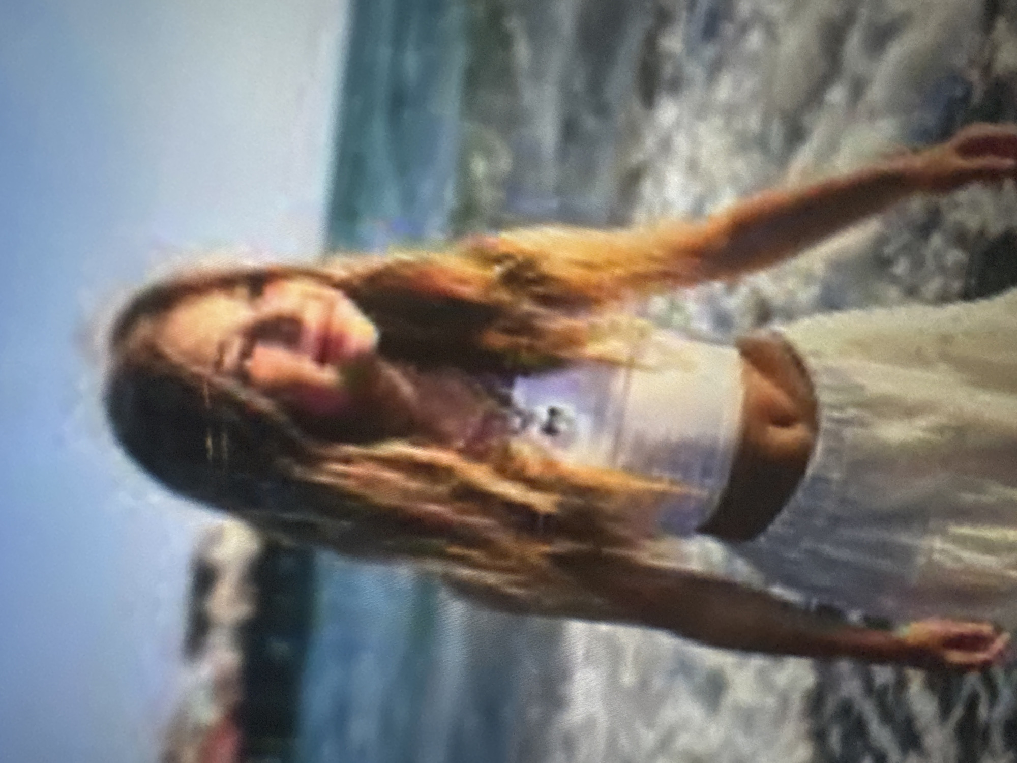 The image displays a young person, likely female, with long, flowing light brown or blonde hair, captured in an outdoor setting by a body of water. The photo is rotated 90 degrees counter-clockwise. She is wearing a white sleeveless crop top and a light-colored, possibly white or cream, flowy skirt. Her midriff is visible. The subject is positioned sideways to the camera but has turned her head to face it, offering a soft, joyful smile. Her arms are outstretched, with one arm clearly visible extending to the right side of the frame.
The background consists of blurred shades of blue and gray, strongly suggesting an ocean or large lake with visible white, foamy waves or surf near the shoreline. The lighting indicates it is daytime, likely sunny, given the brightness. The overall mood conveyed is one of happiness, relaxation, or playfulness by the water. The image quality is somewhat blurry and pixelated. The provided crops offer closer views of the subject's face, hair, and the watery background but do not reveal additional distinct details due to the blurriness. There is no visible text in the image.