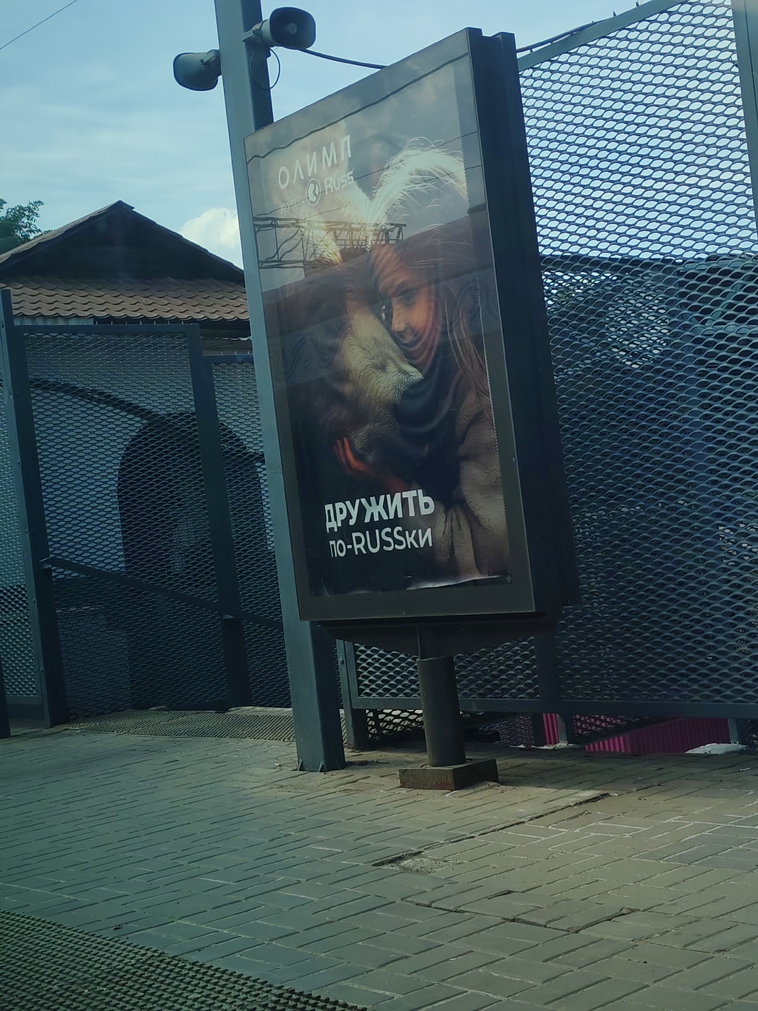 The scene is a public area, most likely a sidewalk, in Moscow, Russia. A billboard stands prominently in the foreground, displaying an advertisement for 'OLIMP' and 'Russ' group, with the slogan "ДРУЖИТЬ по-RUSSKI" (Friendship the Russian Way). The advertisement features an image of a woman and a dog. The billboard is positioned on a grey brick pavement and is attached to a metal pole. In the background, a dark grey metal fence with a diamond-shaped mesh pattern is visible. Behind the fence, a building with a brown tiled roof is discernible. Above the billboard are two loudspeaker horns mounted on the same pole. The overall impression is one of a typical urban environment with commercial advertising.