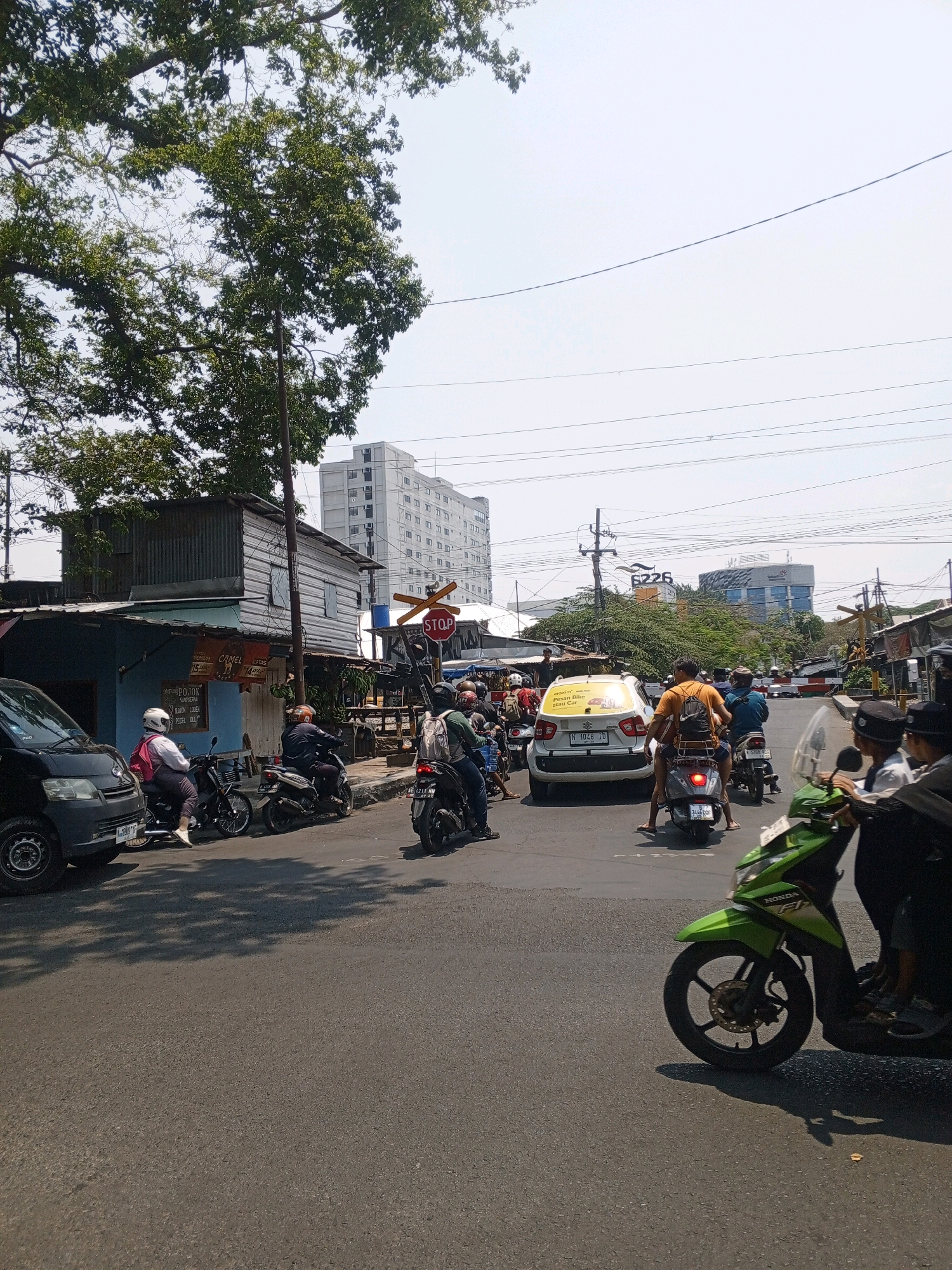 A street scene in Malang, Indonesia features a busy intersection with numerous motorcycles and a few cars. The foreground shows the asphalt road, with shadows of trees cast upon it. A bright green Honda motorcycle is prominently displayed on the right, with its rider partially visible. Further down the road, a white car with a yellow "STOP" sign is at a standstill. Several other motorcycles, occupied by riders wearing helmets, are also present, indicating a traffic jam or waiting for a signal.
On the left side of the image, a dark-colored van is parked, and a person is seated on a motorcycle. Behind the van, a small, weathered building with a corrugated metal roof is visible. Across the street, a multi-story building stands tall against a clear sky. A railway crossing signal with a red "STOP" sign is positioned near the buildings, suggesting the presence of a nearby train track. Power lines crisscross the sky, and lush green trees frame the scene from the top left. The overall atmosphere suggests a typical day in an urban environment, with the characteristic mode of transportation being motorcycles.