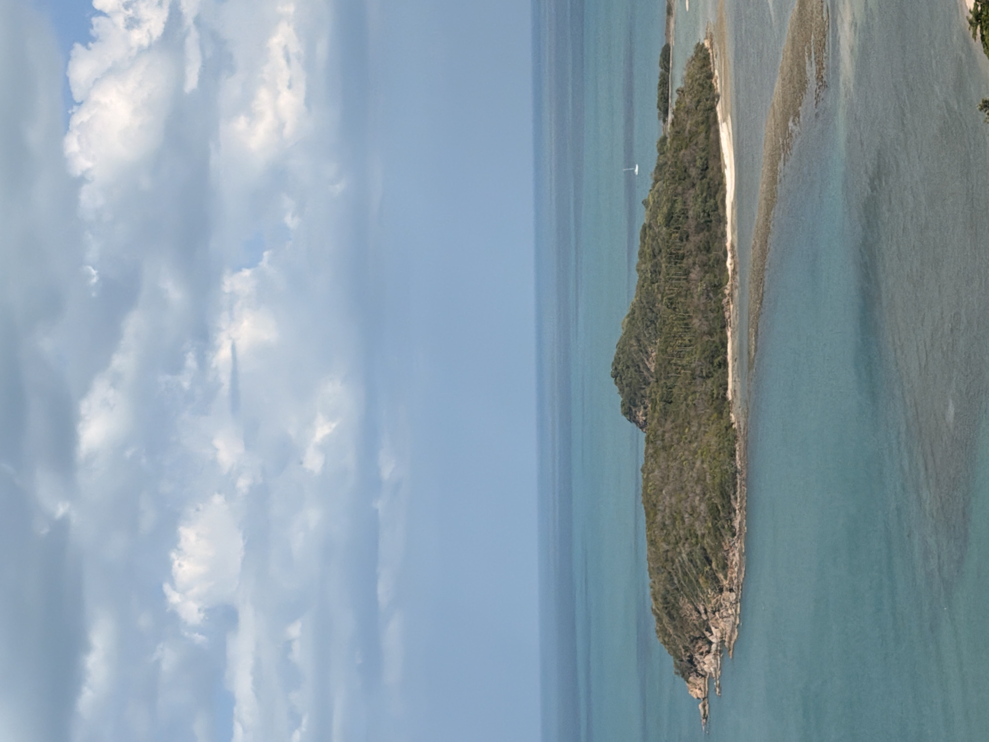 An aerial drone shot captures a lush, green island situated in the turquoise waters of Ko Samui District, Thailand. The island is covered in dense foliage and features rocky outcrops along its coastline. A narrow sandbar connects the main island to a smaller landmass, partially submerged and lined with visible coral formations. The calm, clear water reveals the seafloor in shallower areas. In the distance, a white sailboat glides across the horizon. The sky is a pale blue, adorned with scattered white clouds, some of which appear wispy and thin, while others are more dense and textured. The overall scene is one of serene natural beauty.