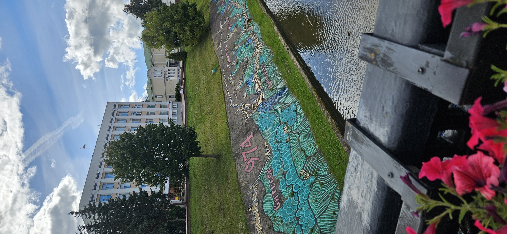 The image depicts a section of a canal in Vitsebsk, Belarus. The canal walls are adorned with a large, colorful mural that appears to depict a map or a series of landmarks, with the number "974" prominently displayed in pink. The mural features stylized waves and buildings, rendered in shades of turquoise, teal, and yellow. The canal itself contains dark, rippling water, reflecting the cloudy sky above.
The canal is bordered by a grassy embankment on the left, which slopes upwards towards a modern, multi-story building with a flag flying from its roof. The building has a pale facade and many windows. To the right of this main building, another, older-style building with a green roof is visible. In the foreground, a dark metal railing runs along the edge of a bridge or walkway, with bright pink petunias in bloom at the bottom of the frame. The sky is partly cloudy, with white clouds drifting across a blue expanse, and a faint contrail from an aircraft is visible. Two individuals are seated on a bench on the elevated area near the buildings, observing the scene.