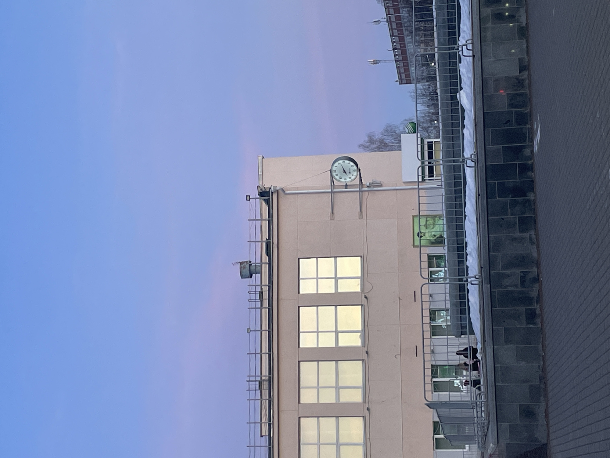 An eye-level, outdoor shot shows a multi-story building in what appears to be the city of Chaikovsky, Russia. The light-colored building has a clock mounted near the top, and multiple windows are lit from within. A metal ladder is affixed to the building's left side. To the right of the building is a paved area. The sky is a blend of light blue and light pink. A metal railing partially obscures the right side of the building and the paved area. Other buildings are visible in the background.