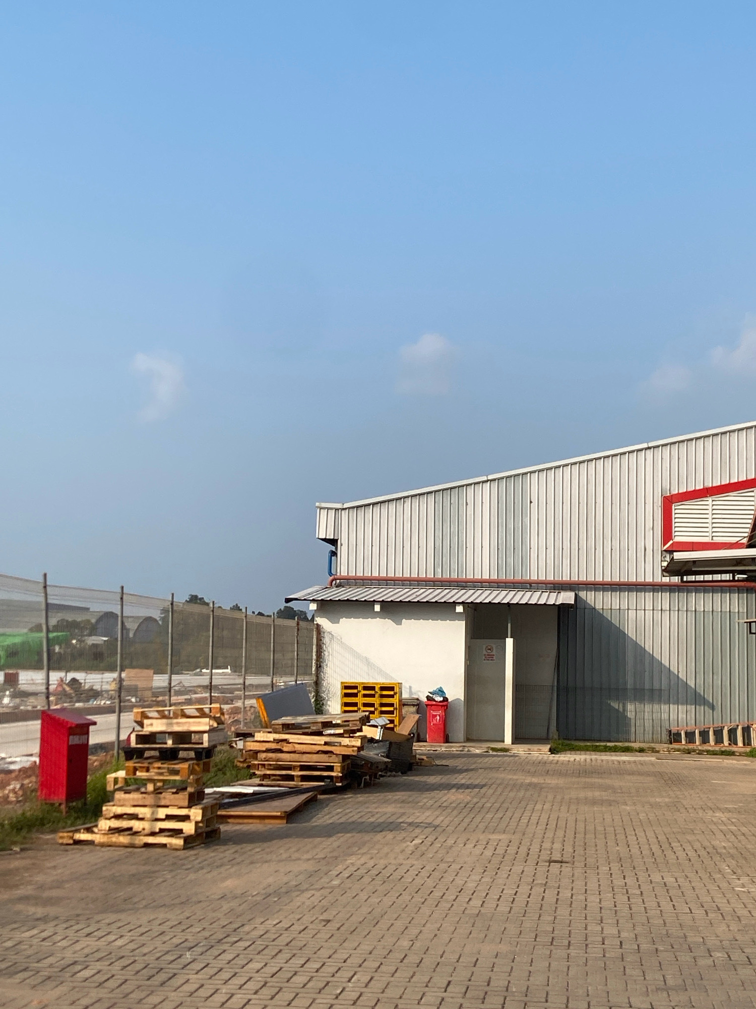 The image shows an outdoor scene in Batam, Indonesia, likely near an industrial or commercial area. In the foreground, a paved surface made of interlocking bricks leads towards a building. To the left, a stack of wooden pallets is piled up, with a red metal bin nearby. Further back, a tall fence encloses an area with some distant structures, possibly greenhouses.
The main subject is a modern building with a corrugated metal exterior and a prominent red and white vent structure on the upper right side. A white section of the building features an open doorway, and a red trash bin and more yellow stacked structures are visible in front of it. To the right of the building, a wooden ladder rests against the wall. The sky is clear blue with a few wispy clouds, suggesting it is daytime. The overall impression is one of a functional, possibly industrial, setting.