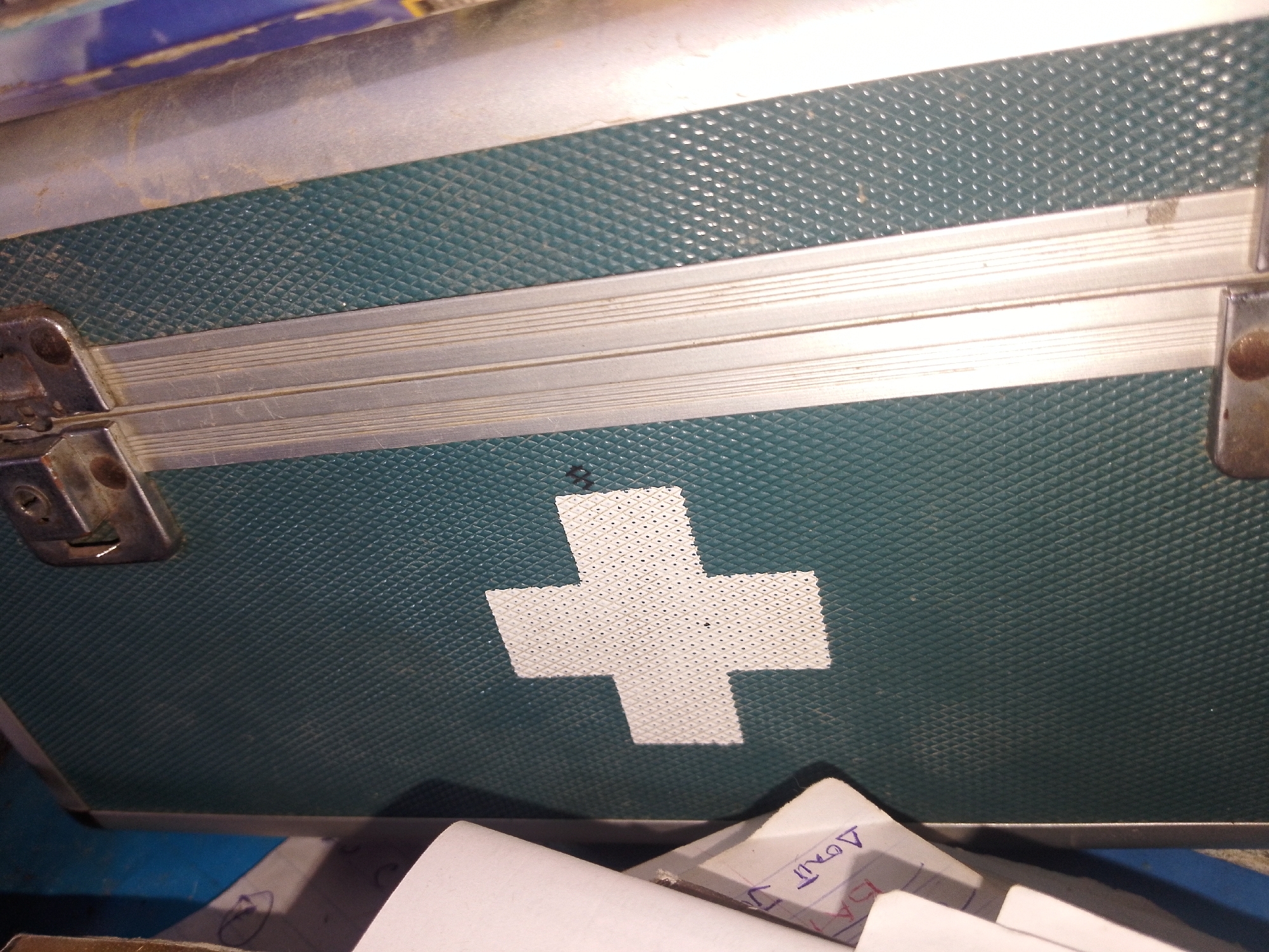 The image is a close-up, high-angle shot of a green medical or first-aid box. The box has a textured surface with a diamond pattern. A white cross symbol is affixed to the front of the box. Silver metal trim runs horizontally across the box, and a silver latch mechanism is visible on the left side. In the lower right and left of the image, parts of white papers with some writing are visible. The scene appears to be indoors, with a blue surface visible beneath the box. There is no visible indication of location within Serti, Nigeria.