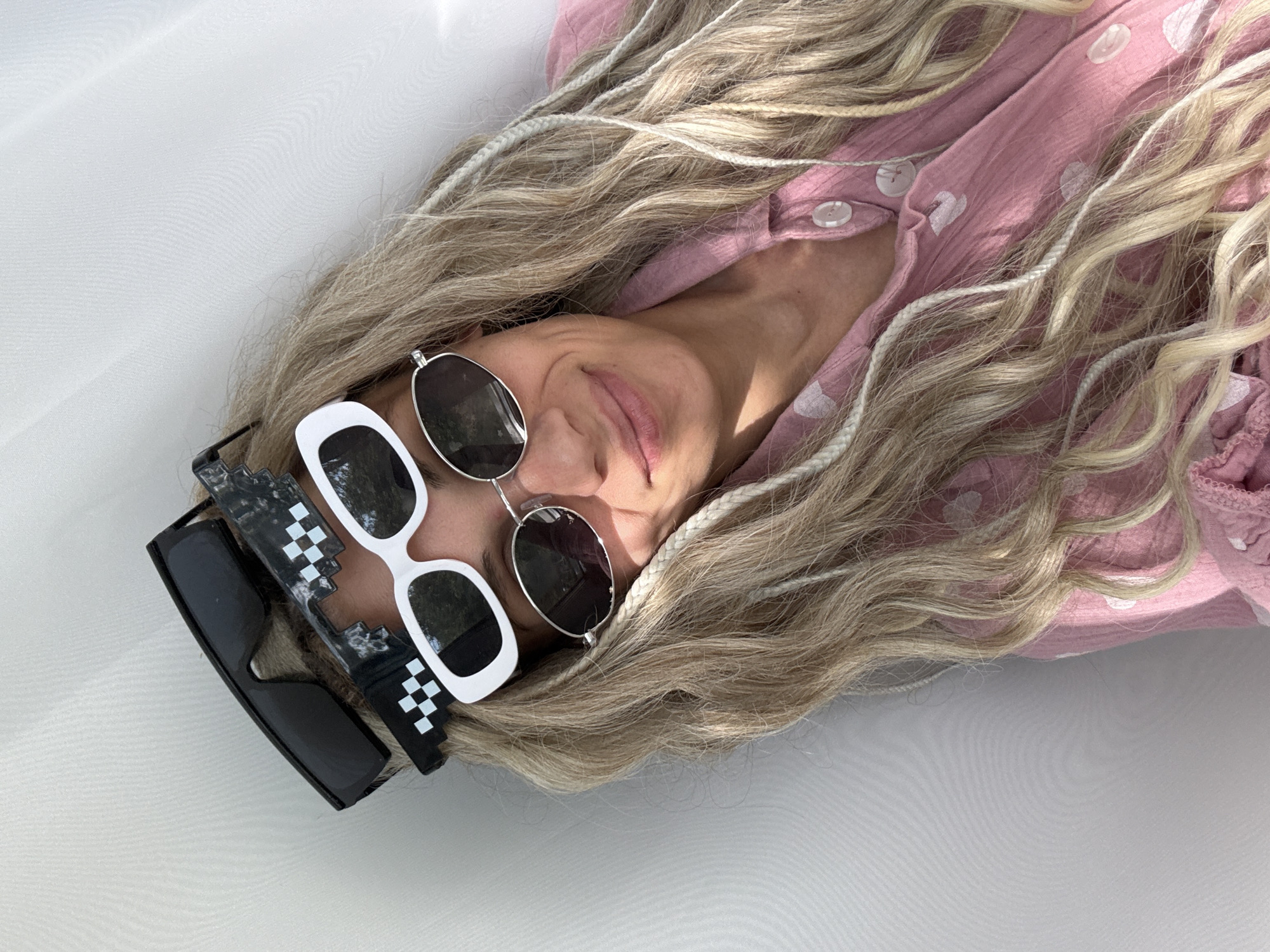 A woman with blonde hair, wearing a pink shirt with white polka dots, lies on a white surface. She is wearing three pairs of sunglasses: one with round, silver frames and dark lenses, one with white frames and dark lenses, and one with black frames and black and white pixelated accents. The scene is well-lit. There is no location information to suggest Rusanovo, Russia.