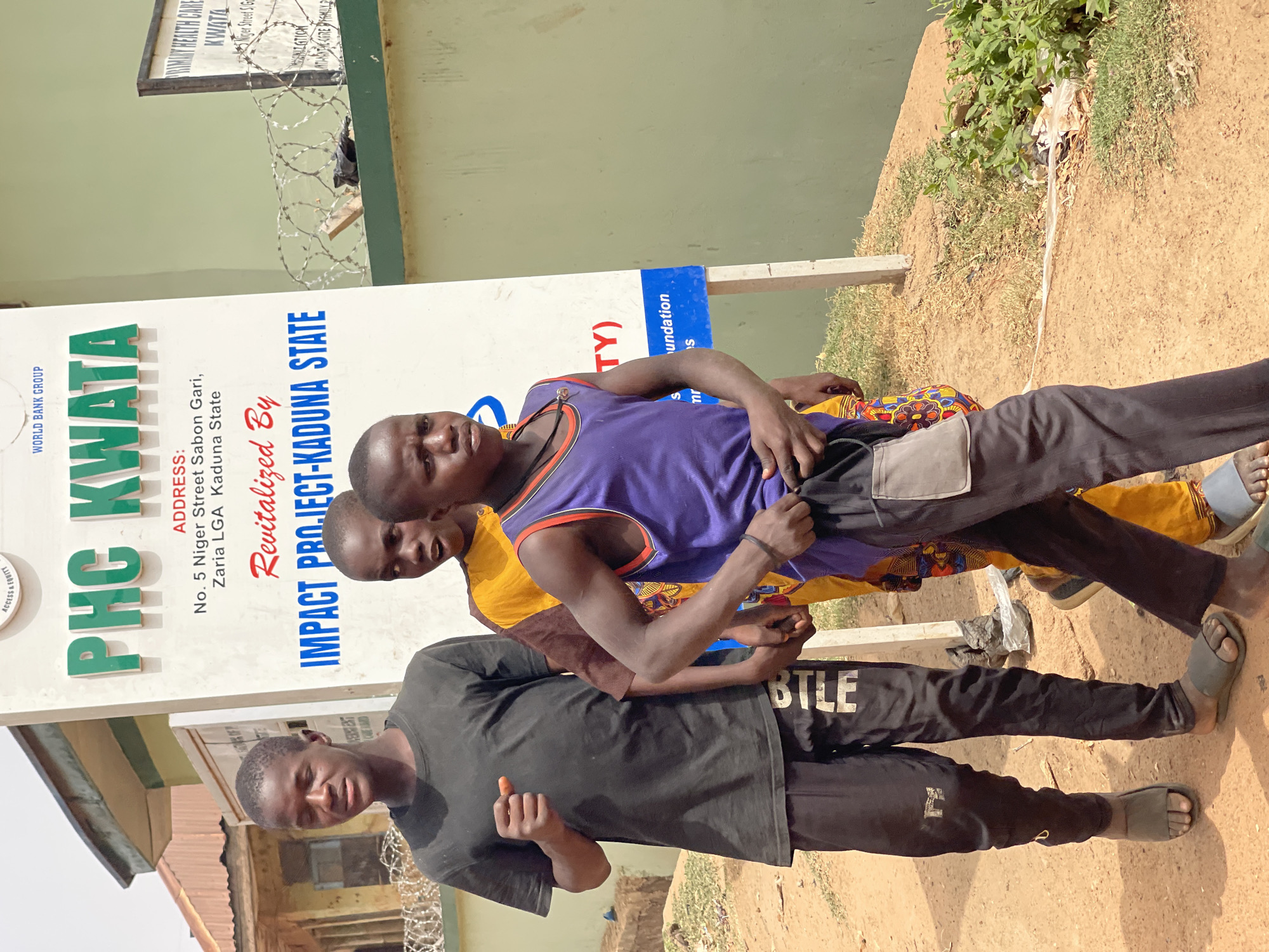 Three young men are captured standing outdoors in Muchiya, Kaduna State, Nigeria, during the daytime under sunny weather. The person in the foreground wears a black t-shirt and black track pants with "BTLE" printed on the side, paired with grey slippers. Behind him, another individual wears a purple and orange sleeveless top, dark trousers, and black sandals. The third person, partially obscured, is dressed in a yellow patterned shirt and dark pants.
They appear to be posing for the camera, looking directly at it with neutral to slightly smiling expressions. Behind them is a large white signpost, indicating the location and purpose. The sign reads "PHC KWATA" (Primary Health Care Kwata) and provides the address: "No. 5 Niger Street Sabon Gari, Zaria LGA Kaduna State." It also states "Revitalized By IMPACT PROJECT-KADUNA STATE" and features the "WORLD BANK GROUP" logo. Other visible text includes "ACCESS & EQUITY." Barbed wire is visible along a fence or wall behind the sign, and the ground is dusty with some sparse vegetation.