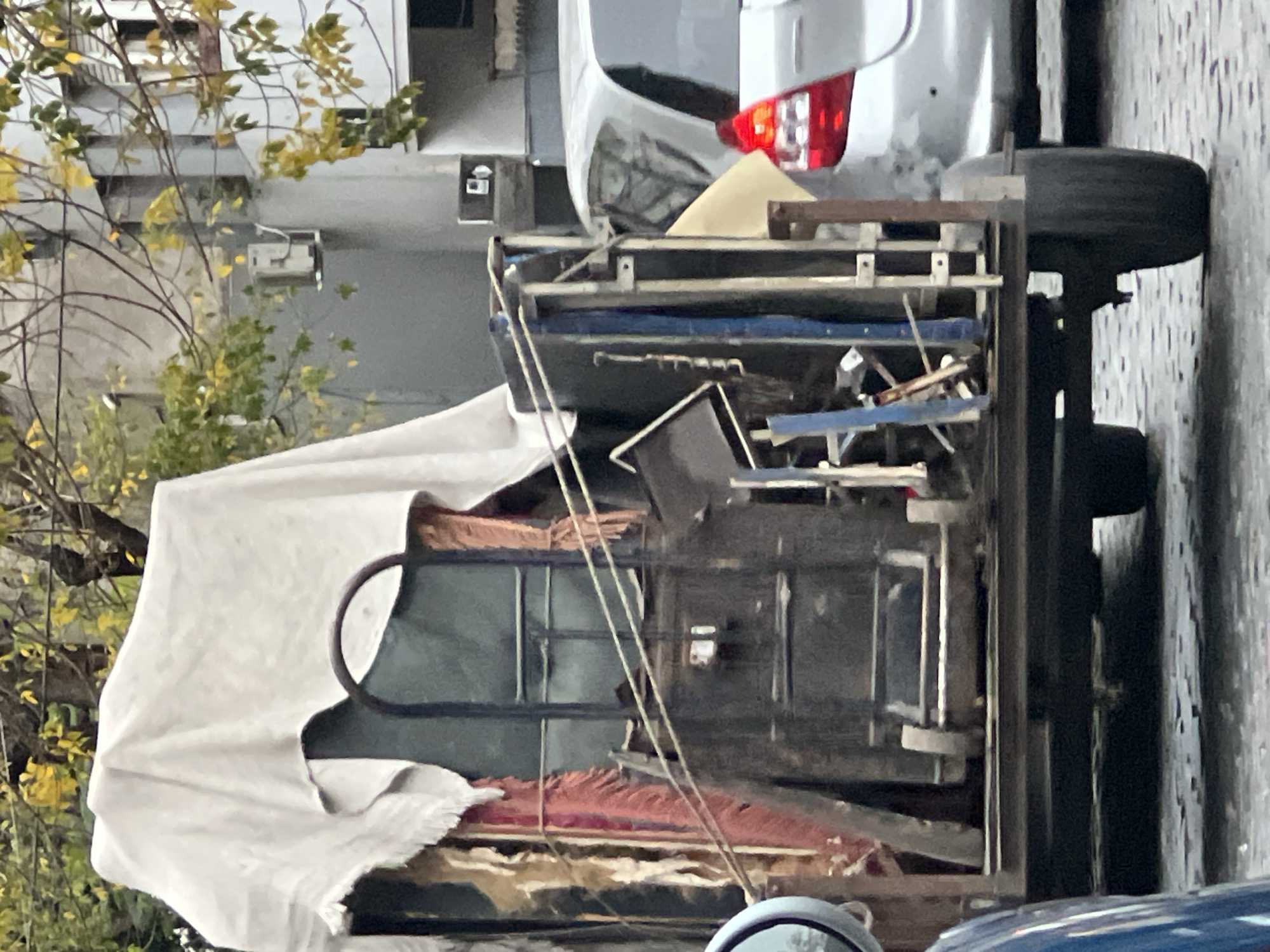 An outdoor photograph shows a vehicle loaded with miscellaneous items, potentially indicating a moving or clean-out situation. The vehicle is partially covered with a white cloth. It appears to be adjacent to a street with a building and trees visible in the background. The red taillight of a car is visible above the load on the trailer, indicating it might be hitched to another vehicle. The location can only be inferred as Buenos Aires, Argentina, based on the prompt's specifications. Further details about the exact street or neighborhood are not discernible from the image.