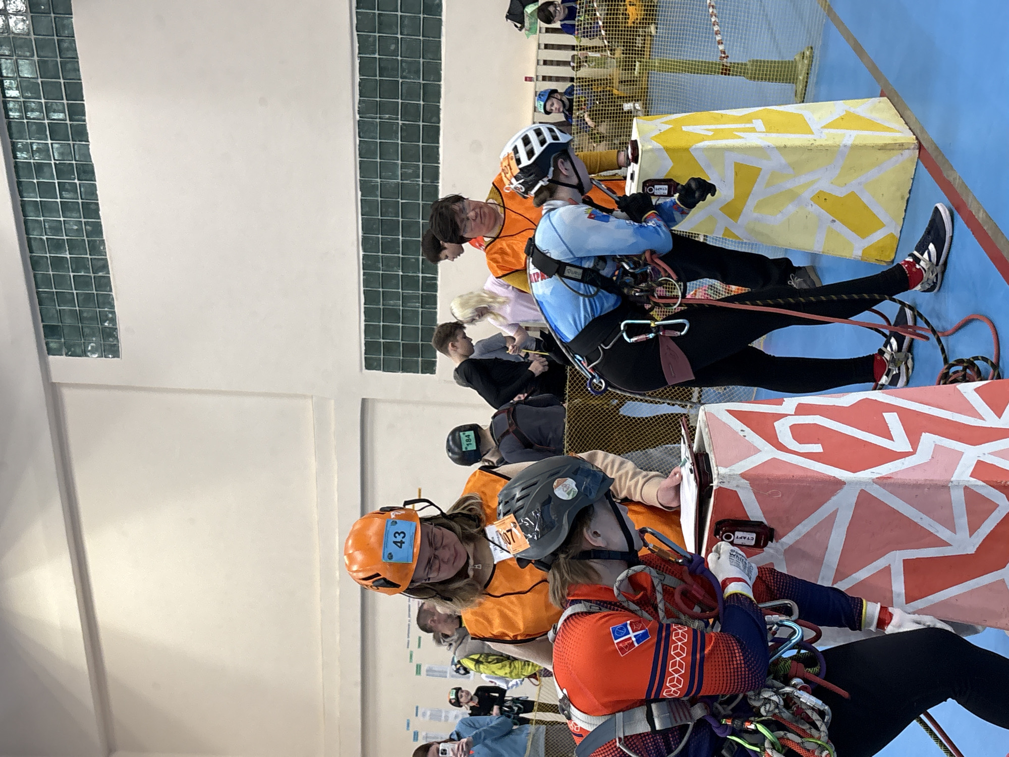 Several people, primarily young adults, are participating in a vertical rope access competition indoors. The setting appears to be a large gymnasium with a blue floor and white walls. Some of the individuals are wearing safety helmets, harnesses, and ropes, and they are connected to structures. They are positioned on or near decorated boxes, which might be part of the competition course. Some participants are observing and taking notes. The competitors seem to be operating equipment mounted on the boxes. The location is Berezniki, Russia.