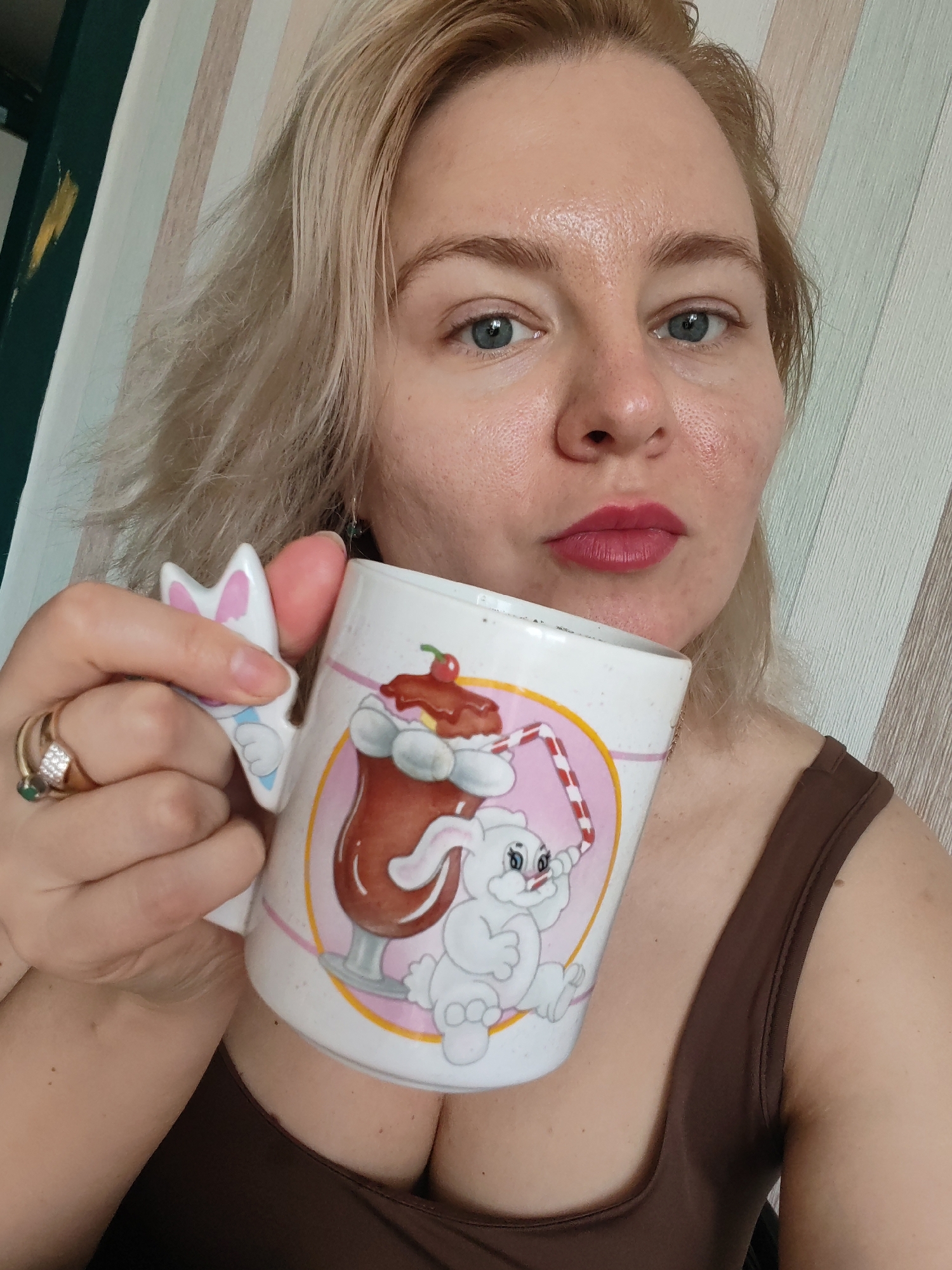 A fair-skinned woman with blonde hair and blue eyes holds a mug with a bunny design in her left hand. She is wearing a brown top. The background features striped wallpaper. The image appears to be taken indoors.