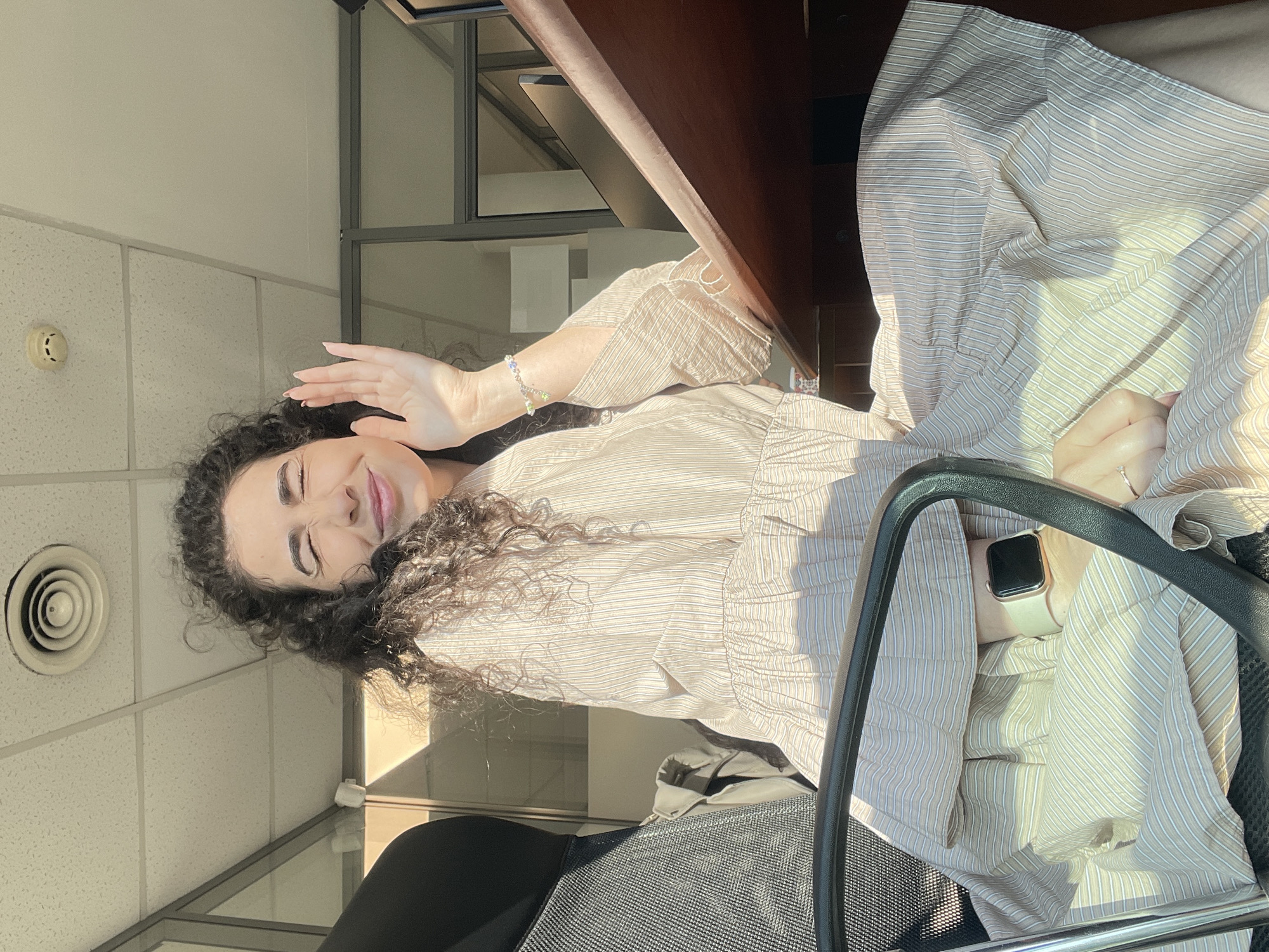 The image shows a woman sitting in an office, with her eyes closed and one hand raised as if waving. She is wearing a striped dress and a watch on her wrist. Sunlight streams in from a window, casting shadows across the room. The office appears to be modern, with glass partitions and fluorescent lighting. The presence of a visible vent on the ceiling and the overall office aesthetic suggest a commercial or professional setting. The location is described as Sofia, Bulgaria.