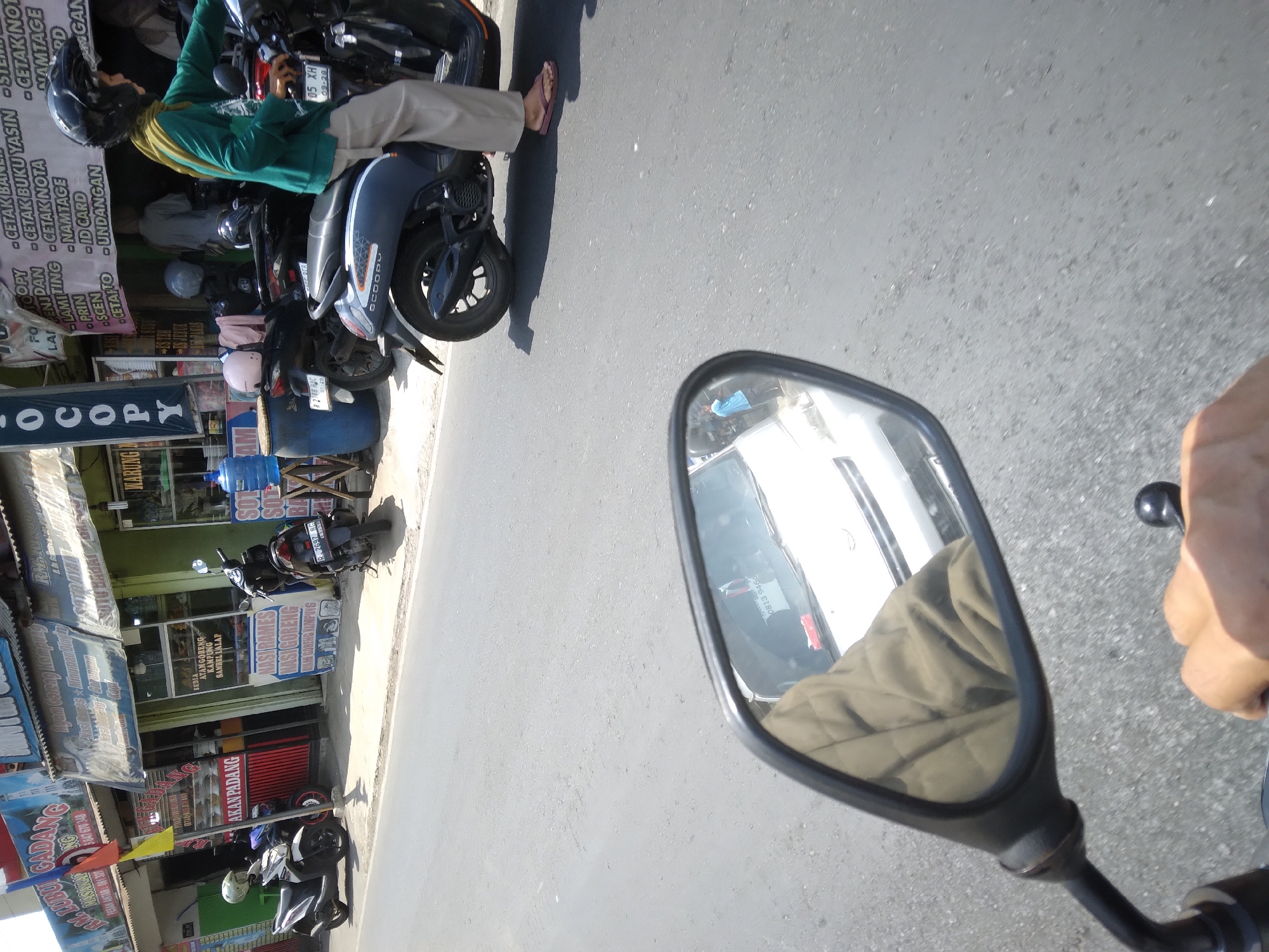 The image depicts a street scene in Desa Pamijen, Kecamatan Sokaraja, Indonesia, on a sunny day. In the foreground, a motorcycle rider's hand is visible on the handlebar, and the motorcycle's side mirror reflects a portion of the street and a white car. In the midground, several parked motorcycles are lined up in front of shops. One of these motorcycles has a rider wearing a green jacket and a black helmet, who is looking to the left. A blue water jug and a dark blue barrel are situated next to the shops. The shops themselves have signs advertising food and services, with one sign reading "AYAM GORENG KAMPUNG SAMBEL TALAP". The background shows more buildings and overhead wires. The overall environment is a busy street with commercial establishments.