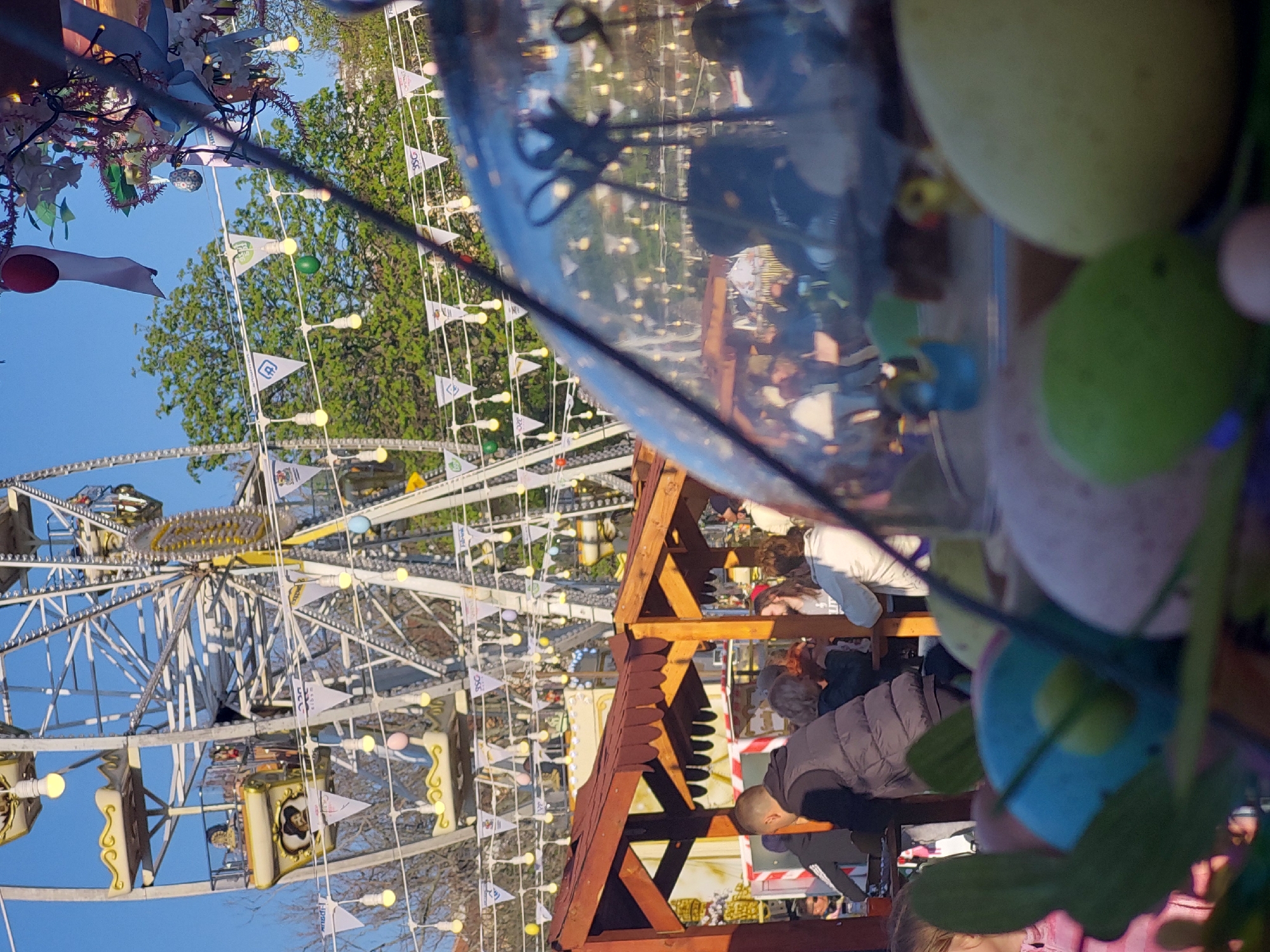 The image captures an outdoor scene at what appears to be a fairground or market, possibly with an Easter theme, under a clear blue sky. A prominent Ferris wheel dominates the left and center background, featuring white metal framework, illuminated light bulbs, and numerous white triangular flags. Several flags bear text such as "DSSD," "DPU," "GSE," and "FRG" or "PRG," along with a symbol resembling a 'B' within a square outline. The Ferris wheel cabins are ornately decorated with gold accents and what appear to be painted portraits.
In the mid-ground, a wooden structure, resembling a stall or pavilion with a pointed roof, is visible, around which several people are gathered. In the right foreground, a large, transparent spherical object acts as a decorative element, containing and surrounded by colorful decorative eggs in shades of green, yellow, pink, and blue, as well as artificial flowers, strongly suggesting an Easter celebration. One person in a dark puffer vest is partially visible near the wooden structure.
The bright natural light indicates it is daytime, likely late afternoon. The overall atmosphere appears casual and festive. A large green-leafed tree is visible in the background behind the Ferris wheel.