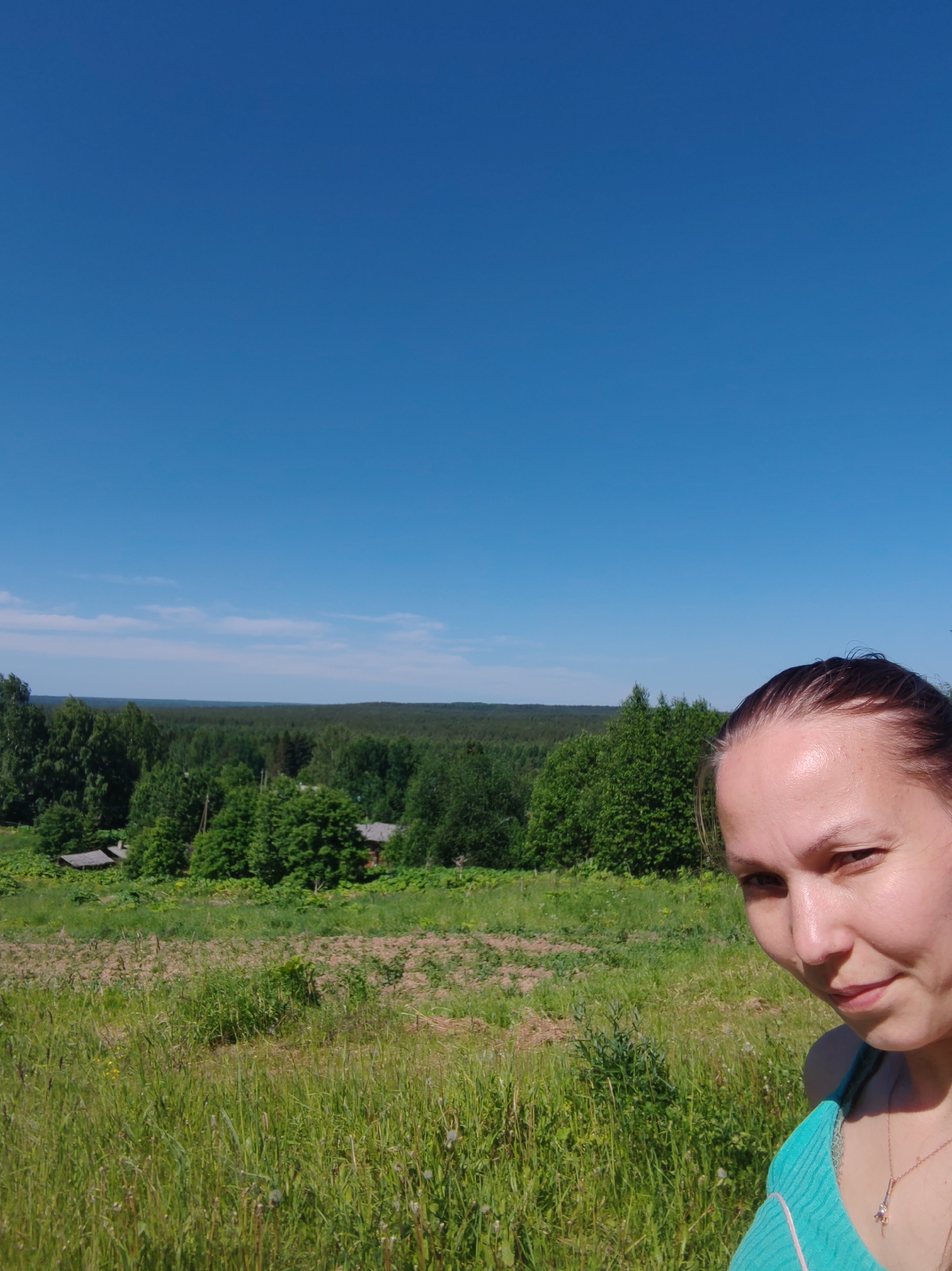 A woman is taking a selfie in a grassy field on a sunny day. She is wearing a turquoise top and a necklace. Behind her, there is a lush green forest and a clear blue sky. The woman's face is partially visible in the bottom right corner of the image. The location appears to be a rural area, likely in Чухлэм, Russia, given the surrounding landscape of forests and fields. The time of day is daytime, with bright sunlight.