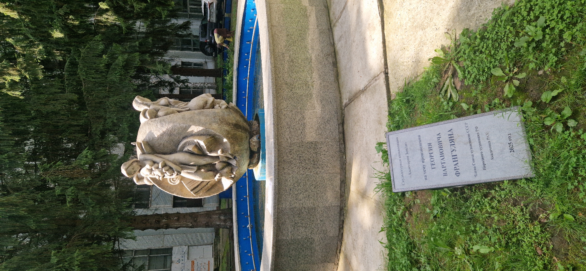 The image shows a public art installation, a fountain with a sculpture, in the city of Krasnoobsk, Russia. The sculpture is made of stone and depicts several figures. It is located in the center of a circular fountain with a blue tiled edge. In front of the fountain, a stone plaque is placed on the grass. The plaque contains text in Russian, which appears to be a dedication to the sculpture and its creator. The background features a building with windows and trees, likely in an urban park or public space. There is no visible action or interaction between people in the scene. The environment appears to be outdoors during the day.