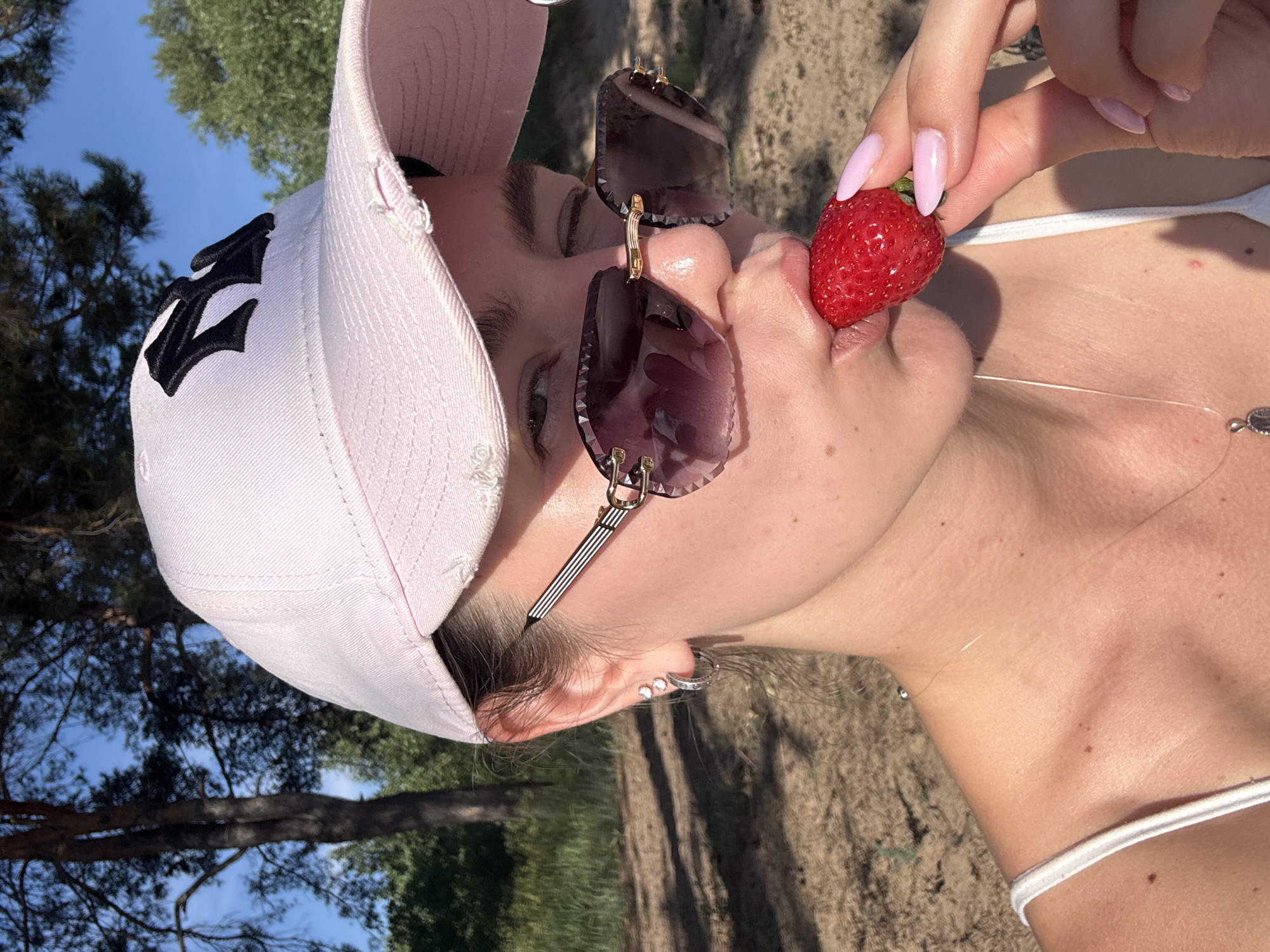 The media file shows a close-up of a person's face and upper body, oriented vertically. The subject is wearing a light pink baseball cap with a black logo on the front. They are also wearing dark-lensed sunglasses with a decorative gold accent. The person is holding a ripe red strawberry with their manicured, light pink fingernails, bringing it towards their mouth. The background consists of greenery from trees and a clear blue sky, suggesting an outdoor setting during the day. The lighting indicates it is daytime, likely sunny. The person's skin is visible, with some freckles on their neck and chest. They are wearing a white top, possibly a tank top or swimsuit, with a thin white strap visible. A delicate necklace is also visible around their neck. The overall mood appears relaxed and summery. There is visible text on the black logo on the cap, but it is not clearly decipherable.