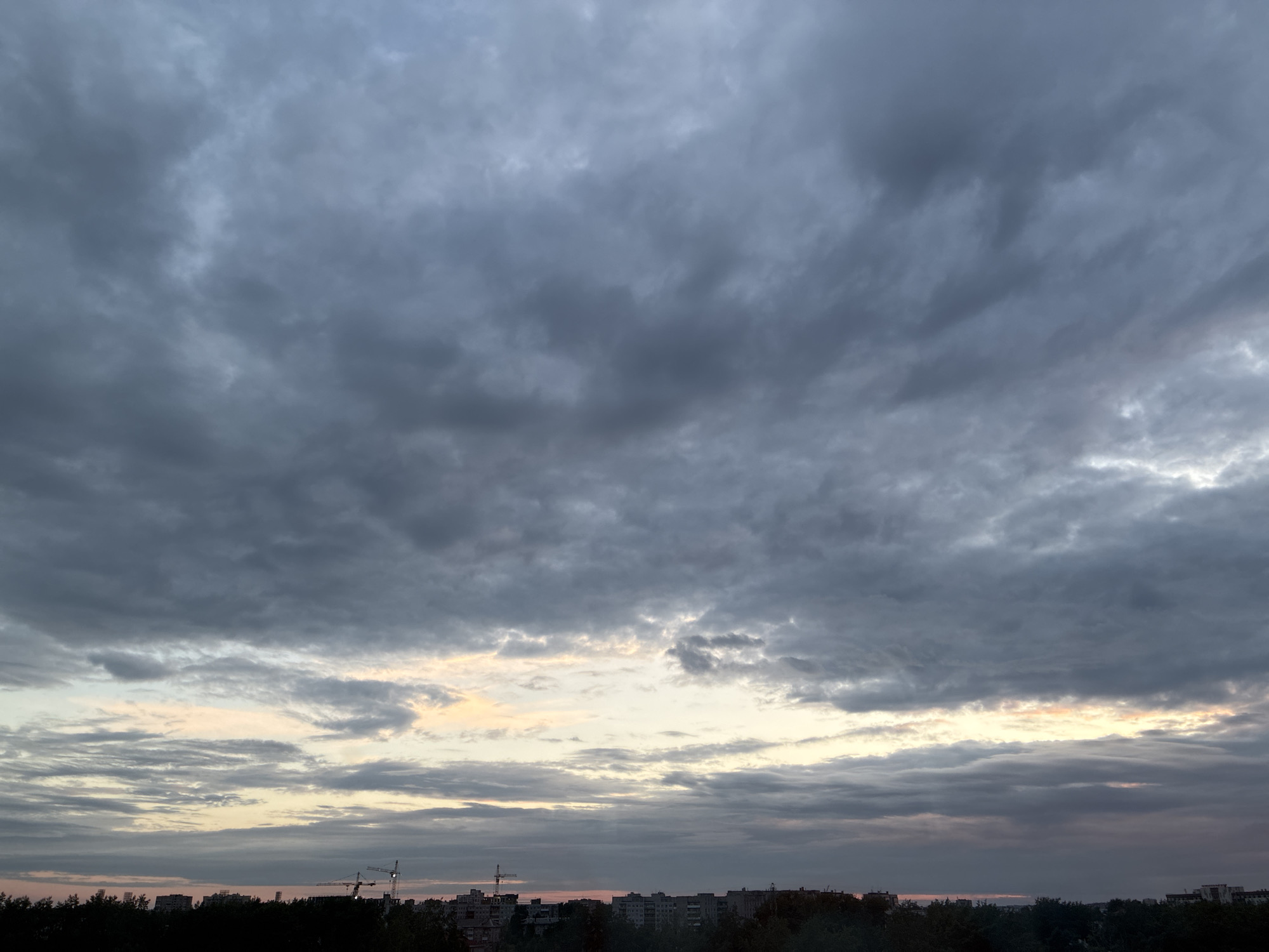 This is a high-angle, full shot of a cloudy sky above a cityscape. The sky is filled with dark, grey, and textured clouds, with a thin band of lighter, yellowish-pink clouds near the horizon. Below the sky, a dark silhouette of trees, buildings, and construction cranes forms the cityscape. The image appears to have been taken at dusk or dawn. There are no people visible in the image. The overall mood is somber and overcast, with a hint of the colors of sunset or sunrise peeking through.