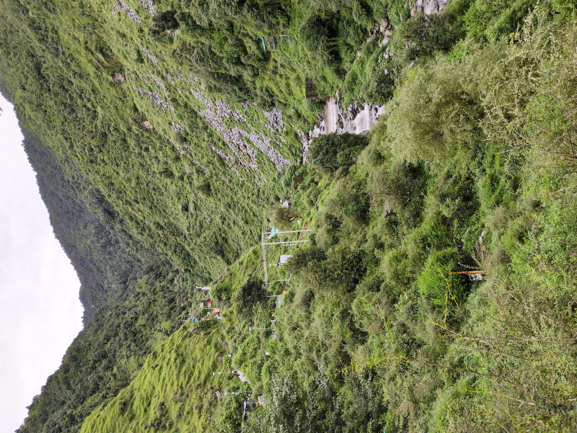 The image depicts a lush, green mountainous valley with a river flowing through it. On the left side of the valley, there are several colorful cabins and a chairlift system ascending the slope. A few people are visible walking along a path. The slopes are densely covered with vegetation, including trees and shrubs. In the upper reaches of the valley, a waterfall can be seen. The river on the right side of the image is rocky and has a small dam or water diversion structure. The sky is overcast. The scene appears to be a natural landscape with some tourist infrastructure. This scene is likely of the Bhagsu waterfall area in Bhagsu, India, known for its scenic beauty and trekking opportunities.