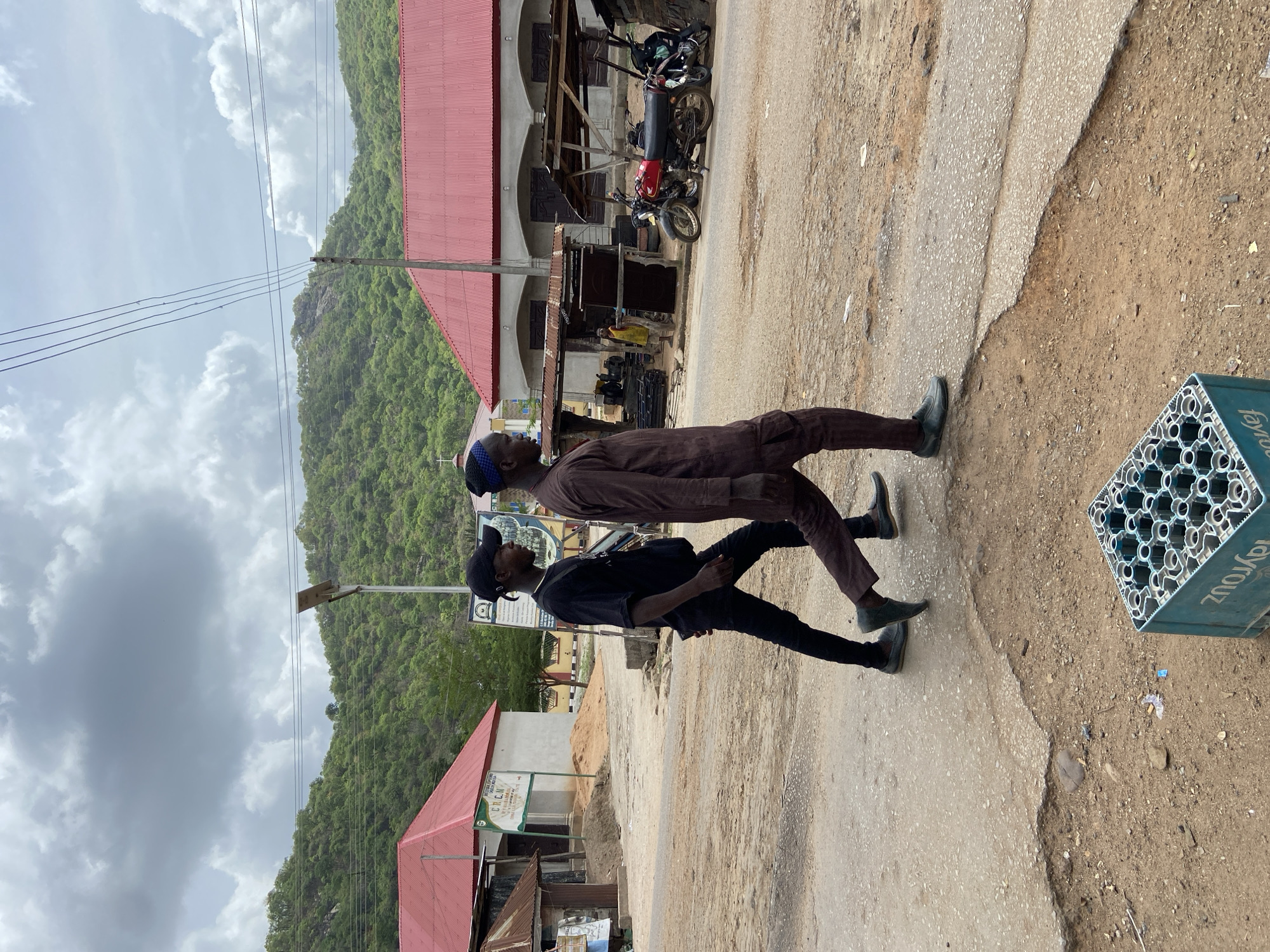 Two men are walking on a road in Mararaba, Nigeria. One man is wearing a brown outfit and a small blue hat. The second man is wearing a black outfit and a black cap. They are walking away from the camera. To the right of the frame is a green plastic beer crate. To the left is a mountain and a cloudy sky. In the background, there are buildings with red roofs, a motorcycle, and a street light.