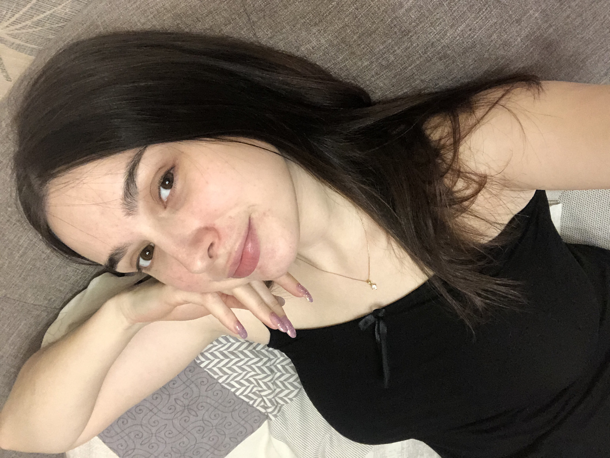 A female individual is depicted in a close-up, top-down perspective within an indoor setting in Chaikovsky, Russia. The subject, a young adult with fair skin and long, straight dark brown hair, lies on her back or side, gazing directly into the camera. She wears a black sleeveless top featuring a small black bow accent near the left shoulder strap, and a delicate gold-colored necklace with a small, light-colored pendant. Her right hand is raised, with fingers positioned near her lower left cheek, showcasing long, manicured nails with a sparkly lavender-pink polish. Her lips are slightly parted, and her dark brown eyes are open. To the left, a patterned fabric, potentially bedding or a pillow, exhibits grey and white geometric designs, including chevrons and a swirling motif. To the right and behind the subject, a textured, plain greyish-brown fabric, consistent with upholstery, is visible. The scene is illuminated by soft, ambient light.