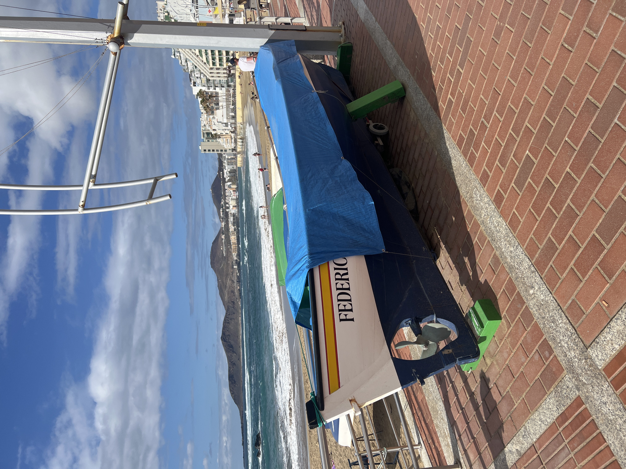 The image captures a vibrant coastal scene in Las Palmas De Gran Canaria, Spain, under bright daylight. In the foreground, a brick promenade, patterned with red bricks and granite accents, hosts two small boats. The nearest boat, named "FEDERICO" in black capital letters on its white, yellow, and blue hull, is covered with a blue tarpaulin and rests on green stands. Its propeller is visible at the stern. A second, similarly covered boat is partially visible behind it.
To the right, a sandy beach unfolds, populated by several individuals enjoying the sun and the ocean. Foamy waves crash against the shore, indicating an active sea. The coastline in the mid-ground is densely lined with white and light-colored city buildings, stretching into the distance. A prominent dark mountain or hill rises in the background, anchoring the urban landscape. The sky above is a clear, vibrant blue dotted with scattered white clouds, contributing to the bright and inviting atmosphere of this sunny day. Metal railings are visible on the left, possibly part of a walkway or pier structure. The scene portrays a typical bustling beach environment within a Spanish island city.