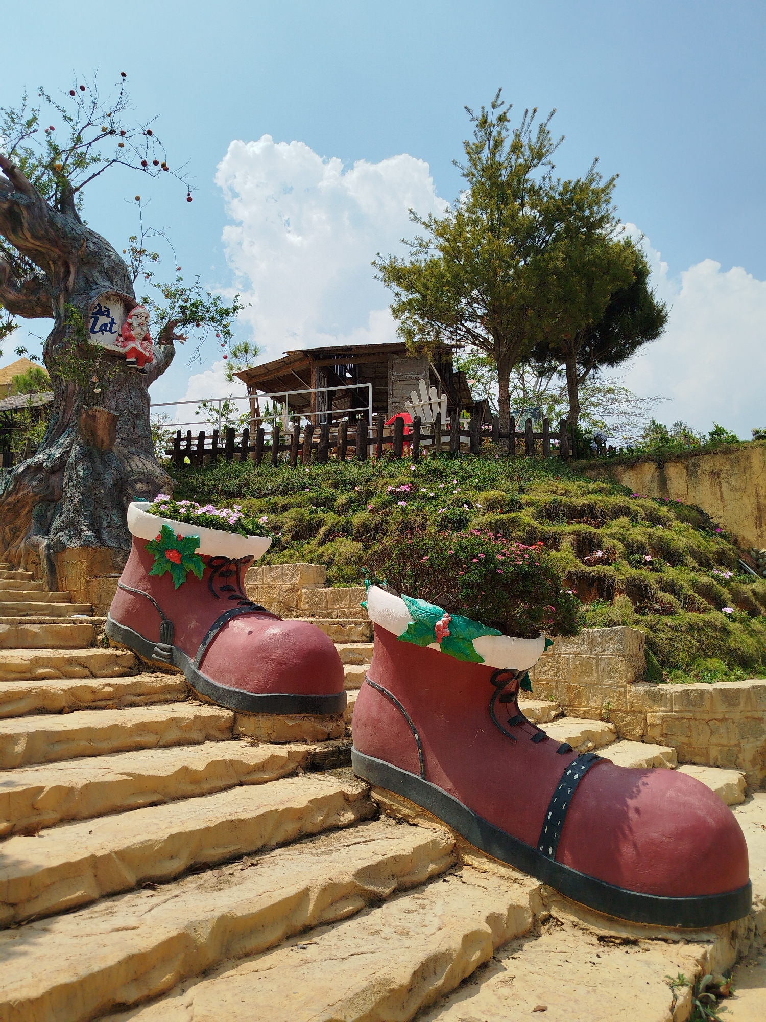 The image captures a sunny outdoor scene, likely in a park or tourist attraction. Two oversized, decorative red boots are positioned on a set of yellow-toned stone steps, filled with plants. The boots have black soles and black "laces" with green and white decorative trim around the top.
Behind the boots, a small wooden structure with a fenced-in area stands atop a grassy hill dotted with pink flowers. A stylized tree sculpture with a sign that appears to read "Dalat" and a Santa Claus figure attached, and mature trees are behind the wooden structure.
The sky is blue with scattered clouds. The image's composition and details suggest the location is Lang Biang - Đà Lạt, Vietnam, based on the distinct architectural and decorative elements.