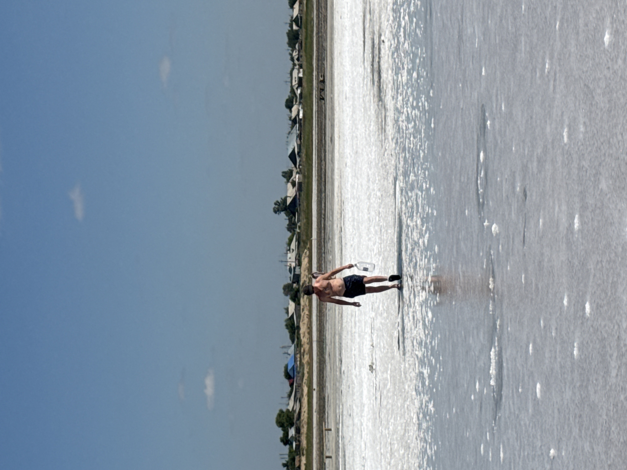 A man, shirtless and wearing dark shorts and a cap, walks across a bright, shallow body of water, likely a salt lake, carrying a large clear plastic bottle. The water's surface is textured with salt deposits, creating a white, crystalline appearance. In the background, a settlement of houses with pitched roofs is visible, nestled amongst sparse vegetation. The sky is a clear, bright blue with a few wispy clouds. The scene is set in Bursol', Russia, a location known for its salt production.