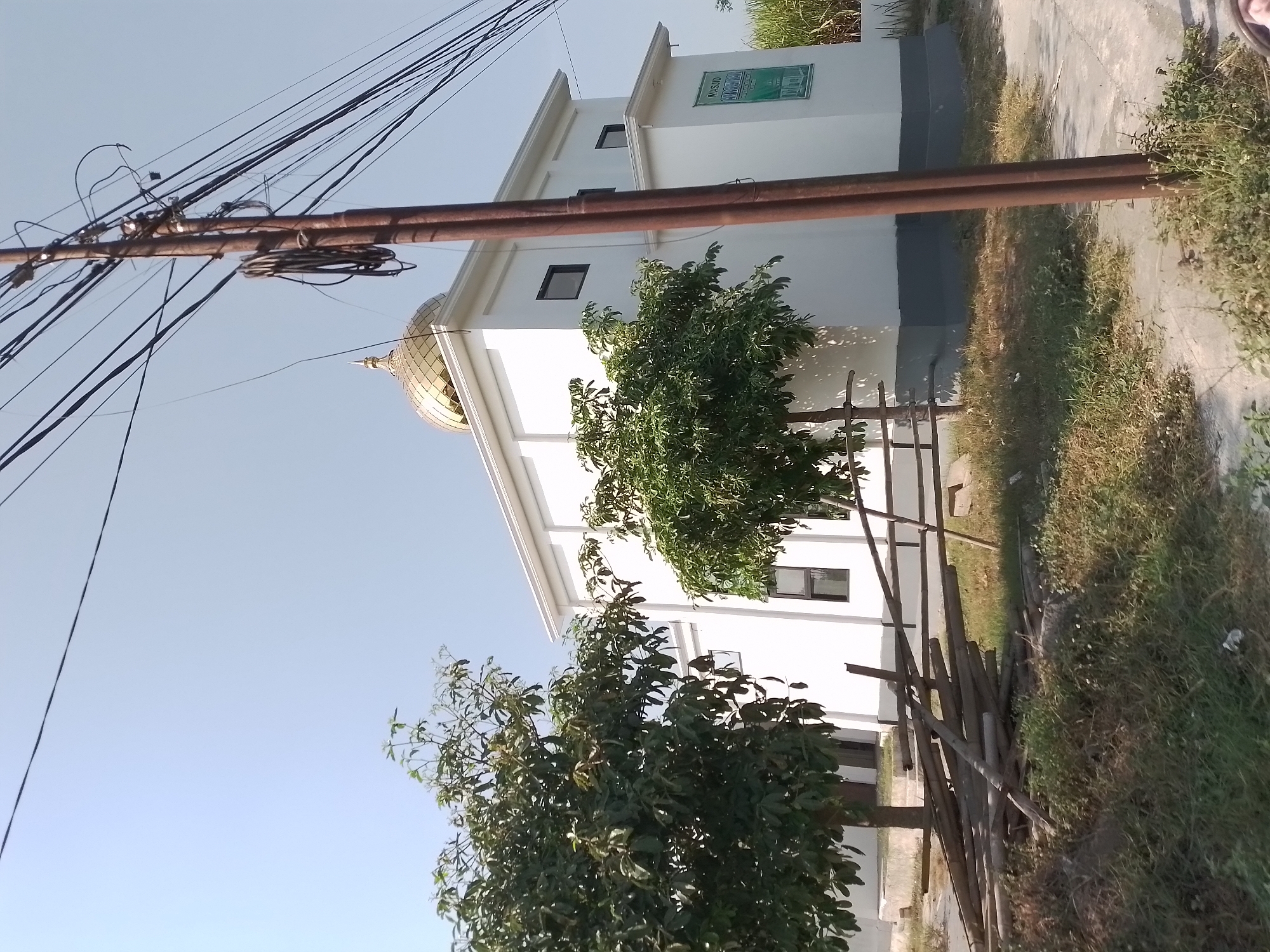 This image depicts a white mosque with a golden dome, located in the city of Ngancar, Indonesia. The mosque is a modern structure with a simple design, featuring dark-framed windows. A green sign with text, likely identifying the mosque, is affixed to the side of the building. In front of the mosque, several trees with lush green foliage are visible. A pile of bamboo poles lies on the ground, suggesting ongoing construction or maintenance. A utility pole with numerous wires stands prominently in the foreground, casting a shadow on the scene. The environment appears to be dry and somewhat unkempt, with patches of grass and scattered debris. The sky is clear and blue, indicating a sunny day.