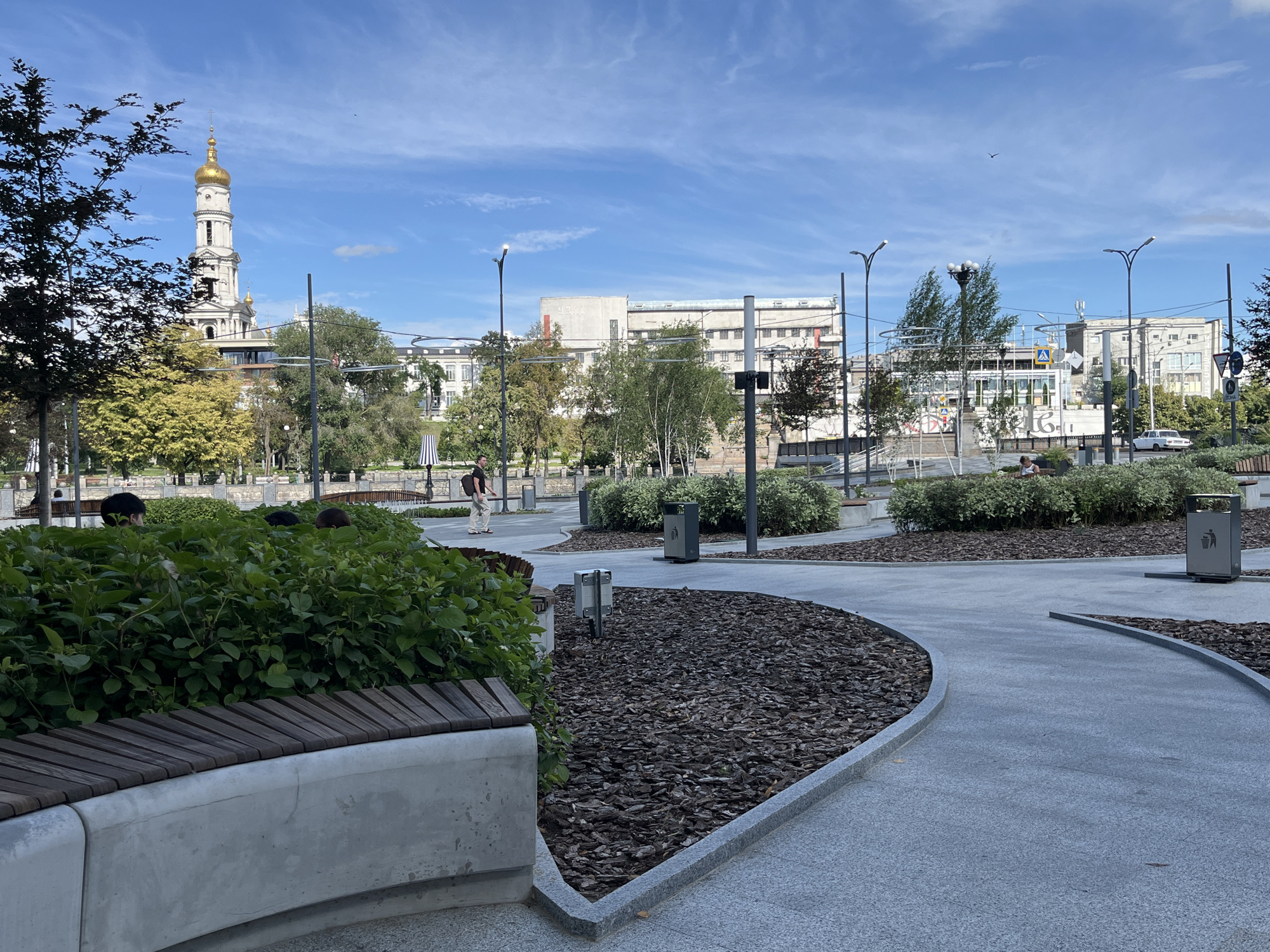 The image shows a public park in Kharkiv, Ukraine, on a sunny day with a blue sky and scattered clouds. In the background, a tall white bell tower with a golden dome, likely part of a church, stands prominently. Several trees with green and yellow foliage are scattered throughout the park. Modern buildings with a mix of architectural styles are visible behind the trees.
In the foreground, a curved pathway made of granite slabs winds through the park. Along the pathway, there are areas covered with wood chips, and modern benches with concrete bases and wooden slats. Green bushes and small trees are planted in these areas.
There are a few people in the park. One person is walking on the pathway in the middle distance, wearing light-colored clothing. In the far background, several figures are seated on benches. Two trash receptacles are visible along the pathway. The scene depicts a well-maintained urban park designed for relaxation and recreation.