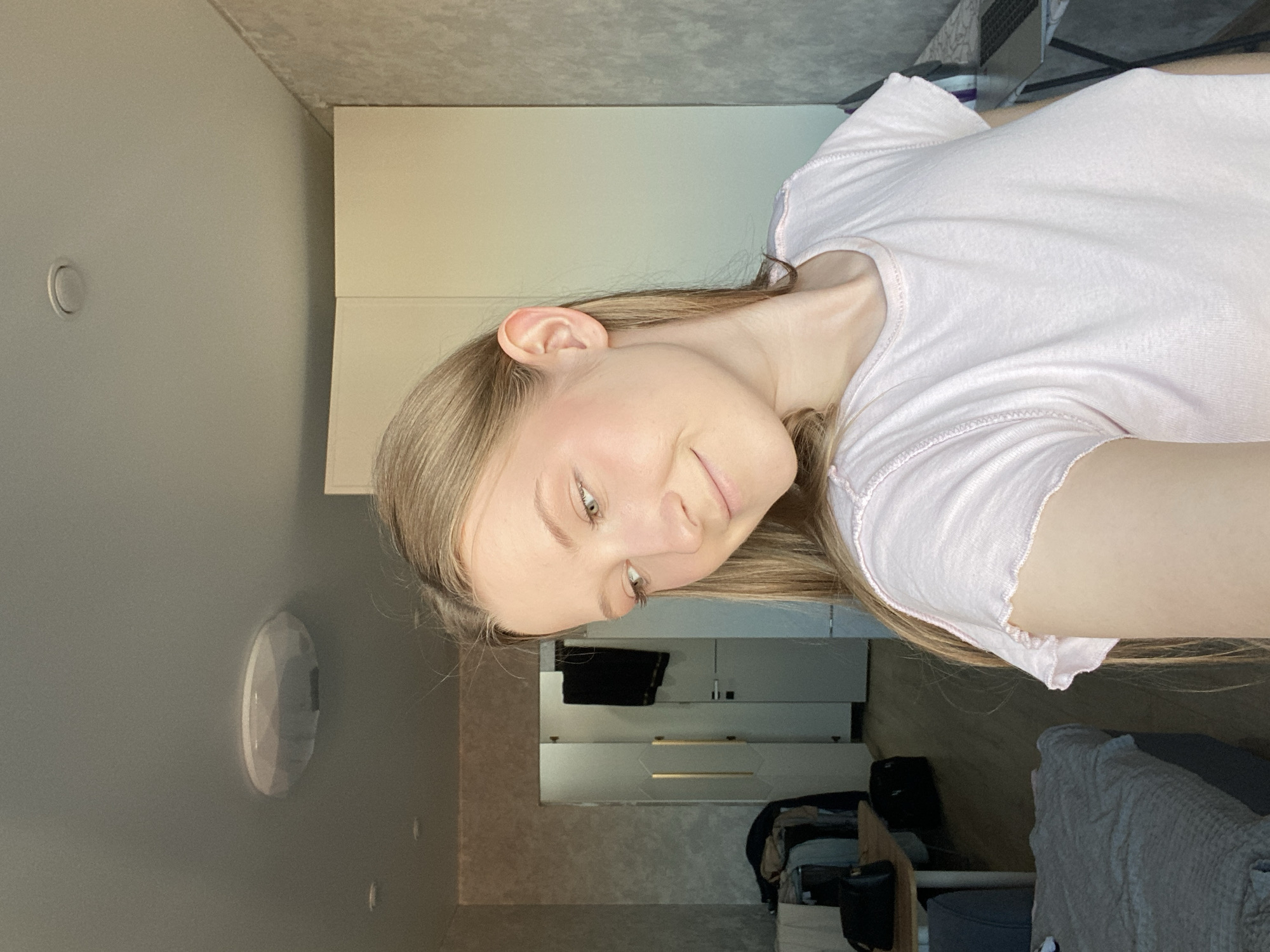 A young woman with long, straight light brown hair and light-colored eyes is captured in a close-up, horizontal portrait, smiling softly. She is wearing a light pink, short-sleeved t-shirt with a ruffled or lettuce-edge trim on the collar and sleeves. Her head is slightly tilted, and her gaze is directed upwards and to the viewer's left, conveying a pleasant and relaxed emotion. Her skin has a fair complexion, and she appears to be wearing minimal makeup, with a hint of blush on her cheeks.
The setting is an indoor room, likely a bedroom or living area, located in Зубово, Russia. The woman appears to be reclining or lying down, with the background objects positioned above and around her. To her left, a light gray wall features a circular recessed light fixture and a white, oval-shaped surface-mounted device, possibly a smart home sensor or smoke detector. Above her, a light-colored, textured ceiling is visible. To her right, a wardrobe or cabinet with light-colored doors stands in the background. Further back and below the wardrobe, shelves or drawers are visible, holding items such as black fabric and small containers. A dark blue or gray upholstered furniture piece, possibly a sofa or bed, covered with a light-colored throw blanket, is also present in the background. The room is brightly and evenly lit, suggesting either natural daylight or well-diffused artificial lighting. There is no visible text in the image.
