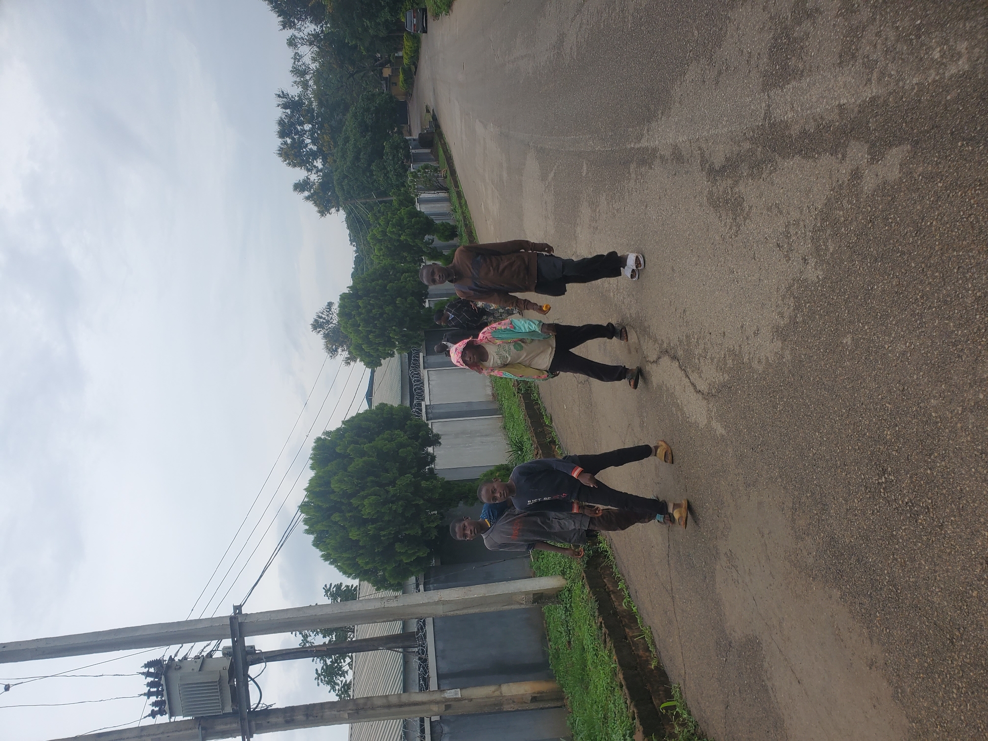 A group of four individuals, appearing to be young males, are walking away from the camera on a paved road in a semi-urban or suburban area. The sky is overcast. The individual on the far left wears a dark gray t-shirt and dark pants, with light-colored sandals. The second individual from the left wears a dark blue long-sleeved shirt and dark pants, also with light-colored sandals. The third individual from the left is wearing a light-colored hooded jacket with colorful patterns and dark pants, and appears to be holding something orange in their right hand. The individual on the far right wears a brown long-sleeved shirt and dark pants, with white sandals.
To the left of the road, there is a curb with overgrown green grass and some bushes. Beyond the grass is a gray concrete wall topped with barbed wire. Behind the wall, there are buildings with corrugated metal roofs and some trees. A utility pole with a transformer and electrical wires stands prominently on the left side of the image.
To the right of the road, the paved surface appears wet in patches. Further down the road, there are more trees and some residential buildings. A dark-colored car is visible in the distance on the right side of the road. The road itself shows some cracks and wear. The scene suggests an everyday street in the city of Jos, Nigeria, characterized by a mix of residential structures, vegetation, and essential utility infrastructure.