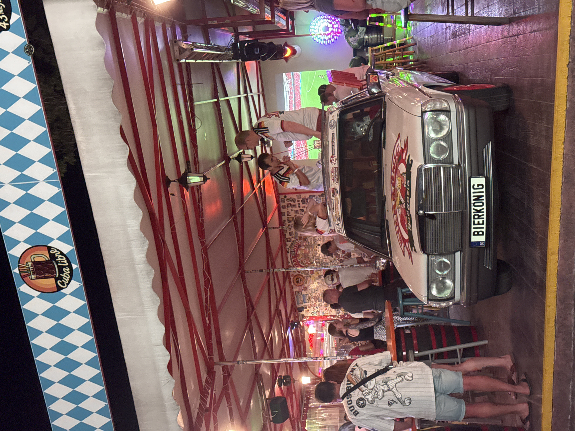 The image depicts an outdoor area in the city of Varna, Bulgaria, likely a bar or entertainment venue, characterized by a festive atmosphere with people gathered. In the foreground, a white Mercedes-Benz car, adorned with stickers and a license plate that reads "BIERKONIG", is positioned prominently. The car appears to be a decorative element or a stage for some of the patrons.
Several people are visible around and on top of the car, some of whom are wearing sports-like jerseys, suggesting a possible event, perhaps a football match, being broadcast on a large screen in the background. One person is standing on the car and drinking through straws. Others are standing or sitting at tables, engaged in conversation and consuming beverages.
The environment is covered by a white and red striped awning, under which various lighting fixtures are installed, creating a colorful ambiance with purple and green neon lights visible. The walls of the venue are decorated with numerous posters and images. The floor is made of wooden planks. In the background, a large screen displays a scene that resembles a football game. The overall scene suggests a lively gathering, possibly celebrating a sporting event in a casual, open-air setting in Varna.