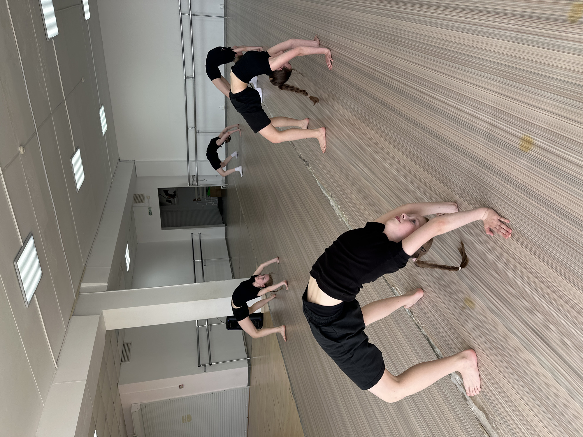 The image shows a dance studio in Votkinsk, Russia. Three female dancers wearing black tops and shorts are performing backbends. The studio has a wooden floor, mirrors on one wall, and ballet barres along another. The room is brightly lit by overhead fluorescent lights. One dancer has a braid.