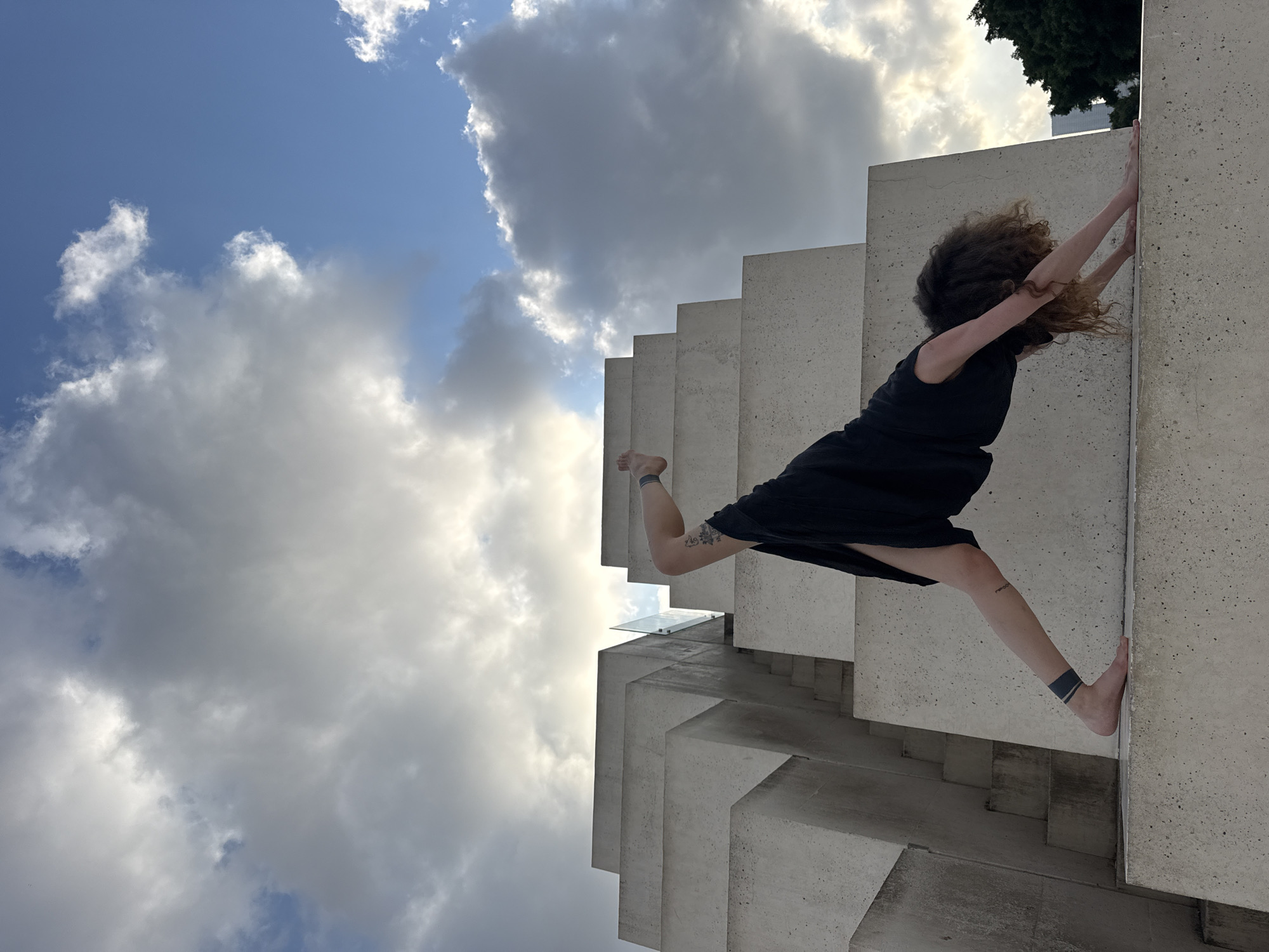 A person is performing a yoga pose on a concrete structure in Tel Aviv, Israel. The structure appears to be made of stacked concrete blocks, forming a stepped or tiered design, with some visible stairs and a glass railing. The sky is cloudy with patches of blue. The person, dressed in black, is in a downward-facing dog pose with one leg extended upward, bent at the knee. Tattoos are visible on the person's leg and ankle. In the background, there is foliage and a building.