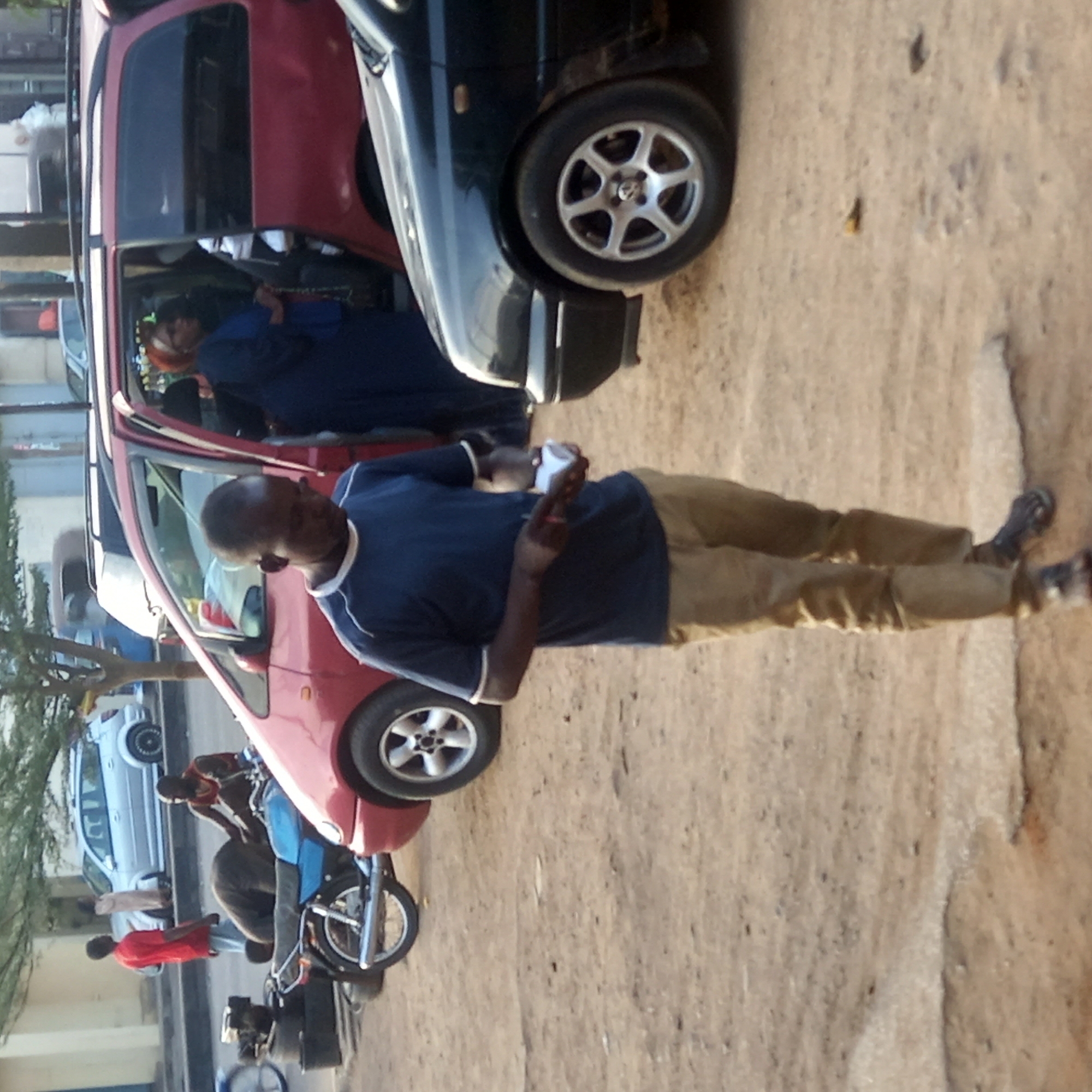 This outdoor street scene, set in Sardauna, Nigeria, captures several people and vehicles on what appears to be an unpaved, dusty road. The image is taken during daylight hours, indicated by the bright, even lighting.
In the foreground, a man of average build stands upright, facing left. He wears a dark blue t-shirt with white trim on the collar and sleeves, and khaki-colored trousers. He holds a white object, possibly a phone or a small cup, in his right hand. His left hand is raised, with fingers partially extended.
Behind him, a red car, possibly a hatchback or compact SUV, is parked with its front passenger door open. Inside the car, a person with reddish hair and dressed in a dark blue garment is visible. Adjacent to the red car, a dark-colored car, also likely an SUV or hatchback, is partially visible, showing its front wheel with silver alloy rims.
Further down the road, two individuals are gathered around a motorcycle. One person, wearing a red shirt, is bending over the motorcycle. Another person, in a blue shirt, appears to be sitting or standing near the motorcycle. Further into the background on the left, a light-colored (possibly silver or white) car is parked. A tree with green foliage is also visible on the left side of the frame, suggesting a roadside setting.
The ground is predominantly unpaved dirt or gravel, with some uneven textures and minor debris visible. There is no visible text in the image. The overall impression is a casual, everyday street scene in a local community.