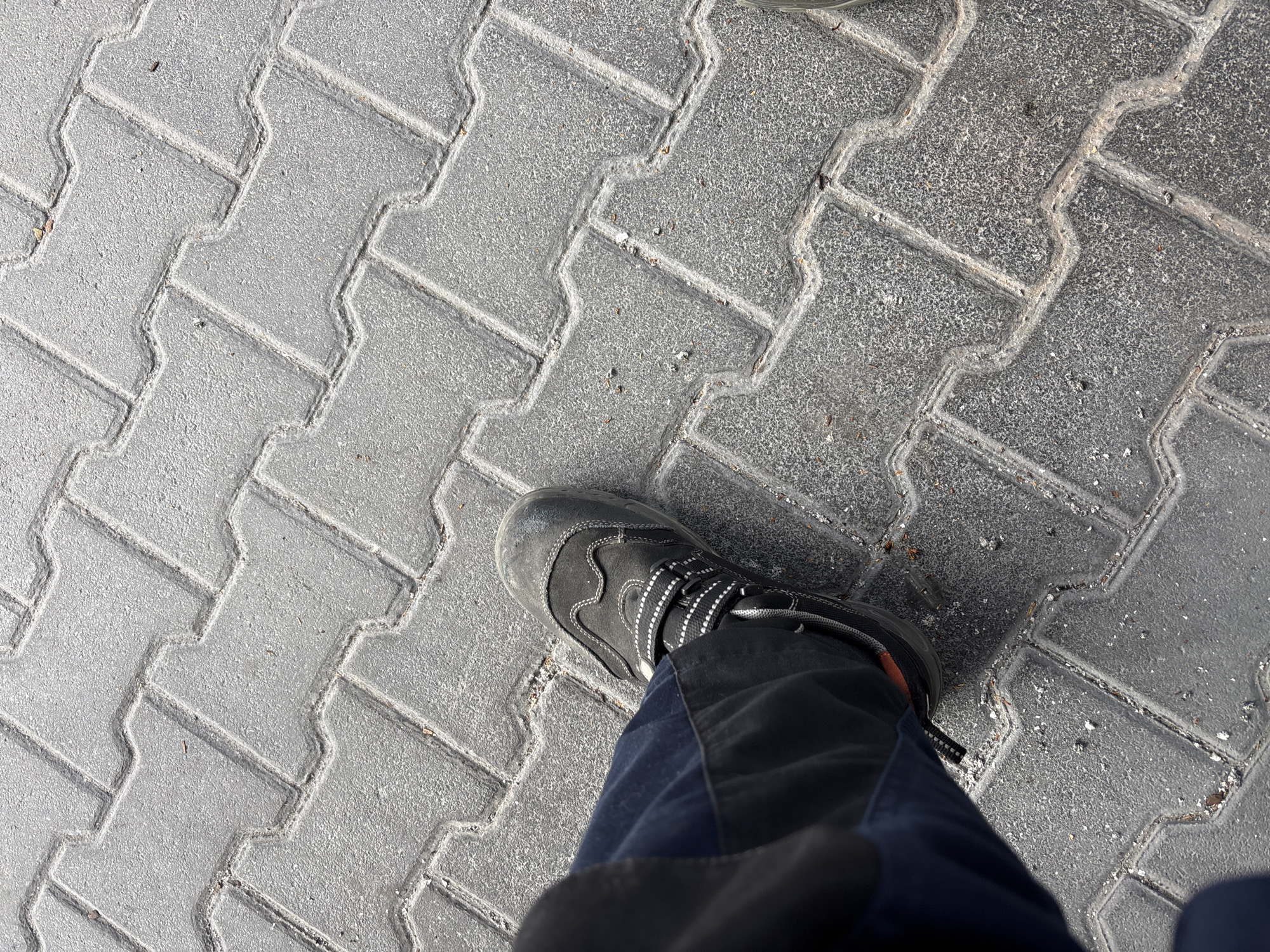 An overhead, high-angle shot shows a person wearing a black work boot and blue work pants standing on a sidewalk made of interlocking grey pavers. The pavers have a textured surface and are laid in a repeating pattern. Some small debris, including small pebbles and organic matter, is visible on the surface of the pavers. The lighting suggests it is daytime, possibly outdoors. The scene appears to be in an urban environment, and specifically, the image was taken at the National Theatre Mannheim, Germany.