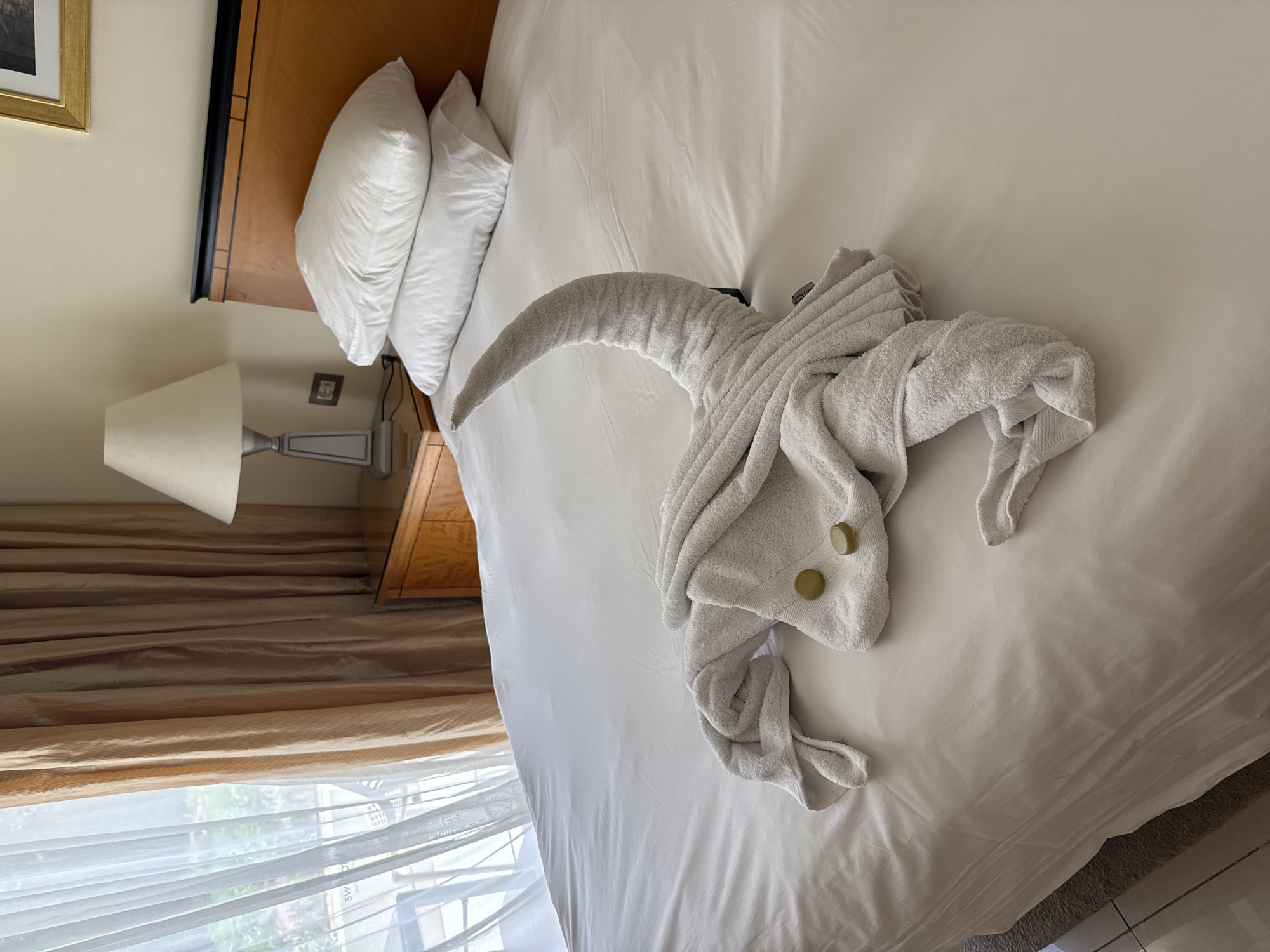 The image depicts a neatly made bed in a hotel room in Sharm El Sheikh, Egypt. The bed is covered with white linens and features two white pillows stacked at the head. On the bed, a white towel is artfully folded to resemble an elephant, complete with a trunk and visible gold-colored buttons as eyes. To the left of the bed, sheer white curtains and heavier beige curtains are drawn, revealing a glimpse of an outdoor area. A wooden nightstand with a glass top sits beside the bed, holding a lamp with a cream-colored shade. The overall scene suggests a comfortable and well-appointed hotel accommodation, possibly in a resort area known for its hospitality services.