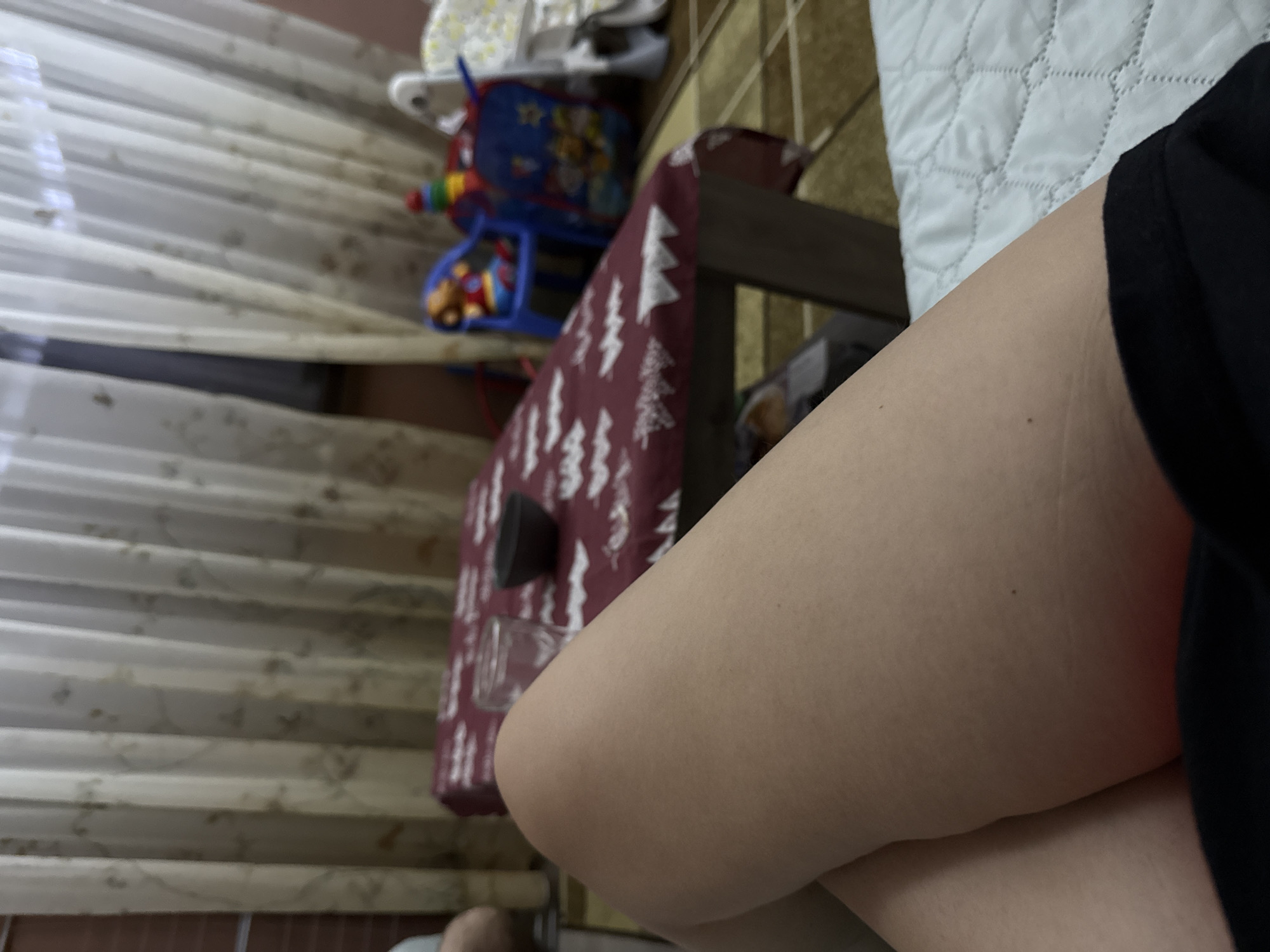The image shows a close-up of a person's leg in the foreground, wearing black clothing, possibly sitting on a bed with a white quilted cover. Behind the leg is a small table covered with a red and white patterned cloth. A black object sits on the table, with a glass container next to it. In the background, there is a curtained window and children's toys. The location is not identifiable as Sofia, Bulgaria from the provided image.