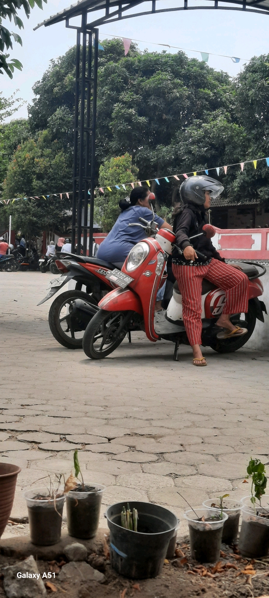 Two individuals are seated on red and white motorbikes in an outdoor, possibly urban, setting. The person in the foreground, a young female wearing a black helmet, a black jacket, and red and white striped pants, is on the rightmost motorbike. She is looking to her right, and her right foot is resting on the ground. The second person, an adult female wearing a blue shirt, is seated on a red motorbike to the left of the first person. She is looking to her left. Both motorbikes have license plates. Behind the motorbikes, there are trees and a structure with a metal frame supporting a roof. Colorful triangular flags are strung across the background. The ground appears to be paved with irregularly shaped stones. In the lower foreground, several potted plants are visible. The image includes a watermark in the bottom left corner that reads "Galaxy A51".