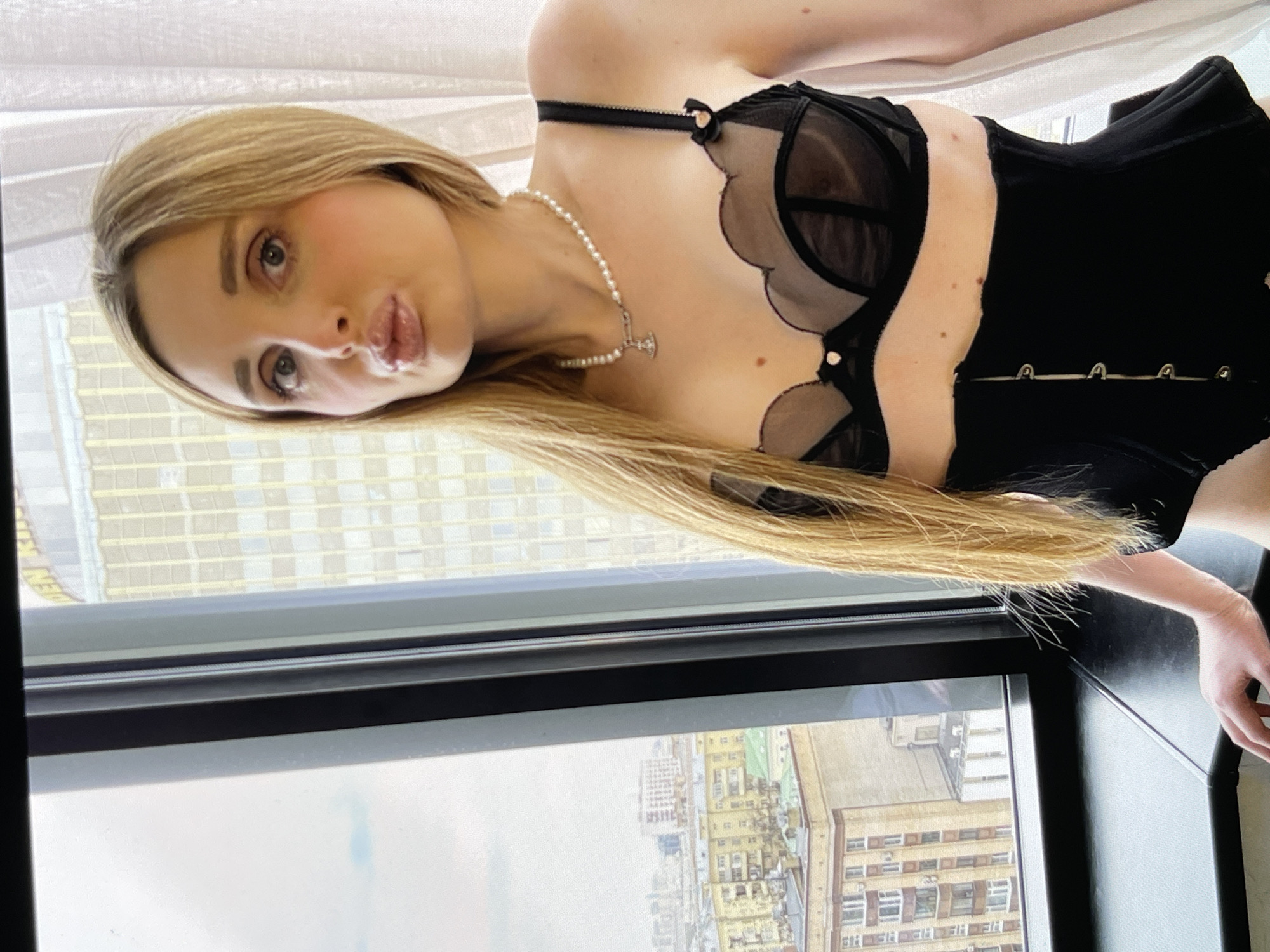 A young woman with blonde hair and light skin is posing in front of a window overlooking a city. She is wearing a black bra and a black corset. The woman is making a kissing face, with her lips puckered. Her hair is long and straight, and it falls down her back. She is also wearing a pearl necklace with a small pendant. The window shows a cityscape with buildings and a cloudy sky. The overall impression is that of a self-portrait taken by the woman.