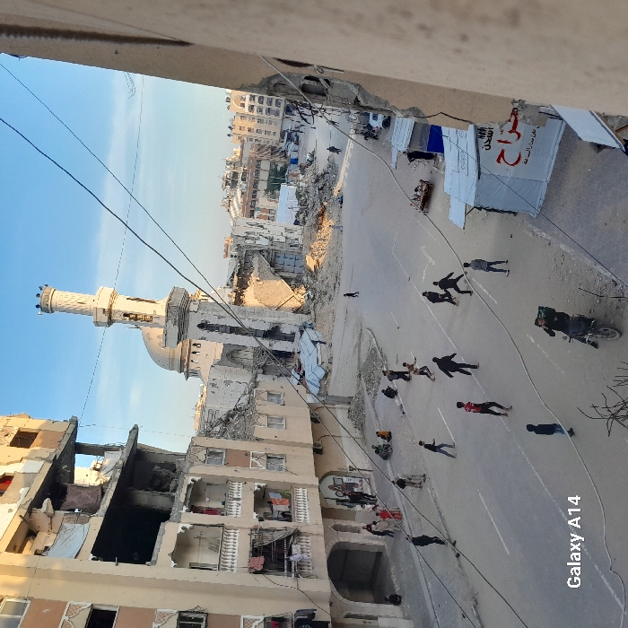 The image captures a street scene in Khan Yunis, Palestinian Territory, during daylight hours under a clear blue sky. The environment is characterized by widespread structural damage to multi-story concrete buildings.
A prominent mosque, featuring a largely intact minaret and dome, stands in the midground, but its lower sections are extensively damaged with significant rubble accumulation around its base. Multi-story residential buildings flanking the left side of the street exhibit severe structural compromise, with visible internal areas, collapsed sections, and debris hanging from balconies. Buildings in the background also show signs of damage.
Approximately 15 to 20 individuals are present on the asphalt street. A cluster of young males, positioned across the street, are engaged in activity suggestive of playing football. A smaller group of people, including adults and children, is gathered on the left sidewalk. One person is seated on a motorcycle in the lower right foreground, facing the camera. Another individual is walking alone further down the street toward the mosque.
A makeshift stall, constructed from white tarps and featuring Arabic text, is visible on the right side of the street. A sofa is placed on the roadside nearby. Overhead utility lines crisscross the upper portion of the frame. Scattered construction debris and rubble are present, particularly around the damaged buildings.