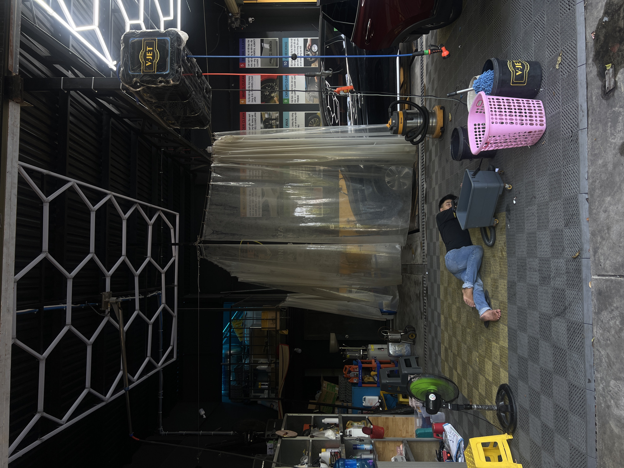 The image depicts an indoor car wash service in Nam Nha Trang Ward, Vietnam. Inside, a worker in jeans and a black shirt, barefoot, is positioned next to a gray container on wheels. Beside him are cleaning supplies including a bucket with "V-JET" printed on it, and a pink basket. Cars undergoing washing are separated by a semi-transparent plastic curtain. A V-Jet branded machine is mounted to the ceiling. A fan is placed near the worker and cleaning supply storage area. The flooring is made of interlocking gray rubber tiles. Signs detailing the car wash process are attached to the wall. A partial view of a car is visible above the curtain.
